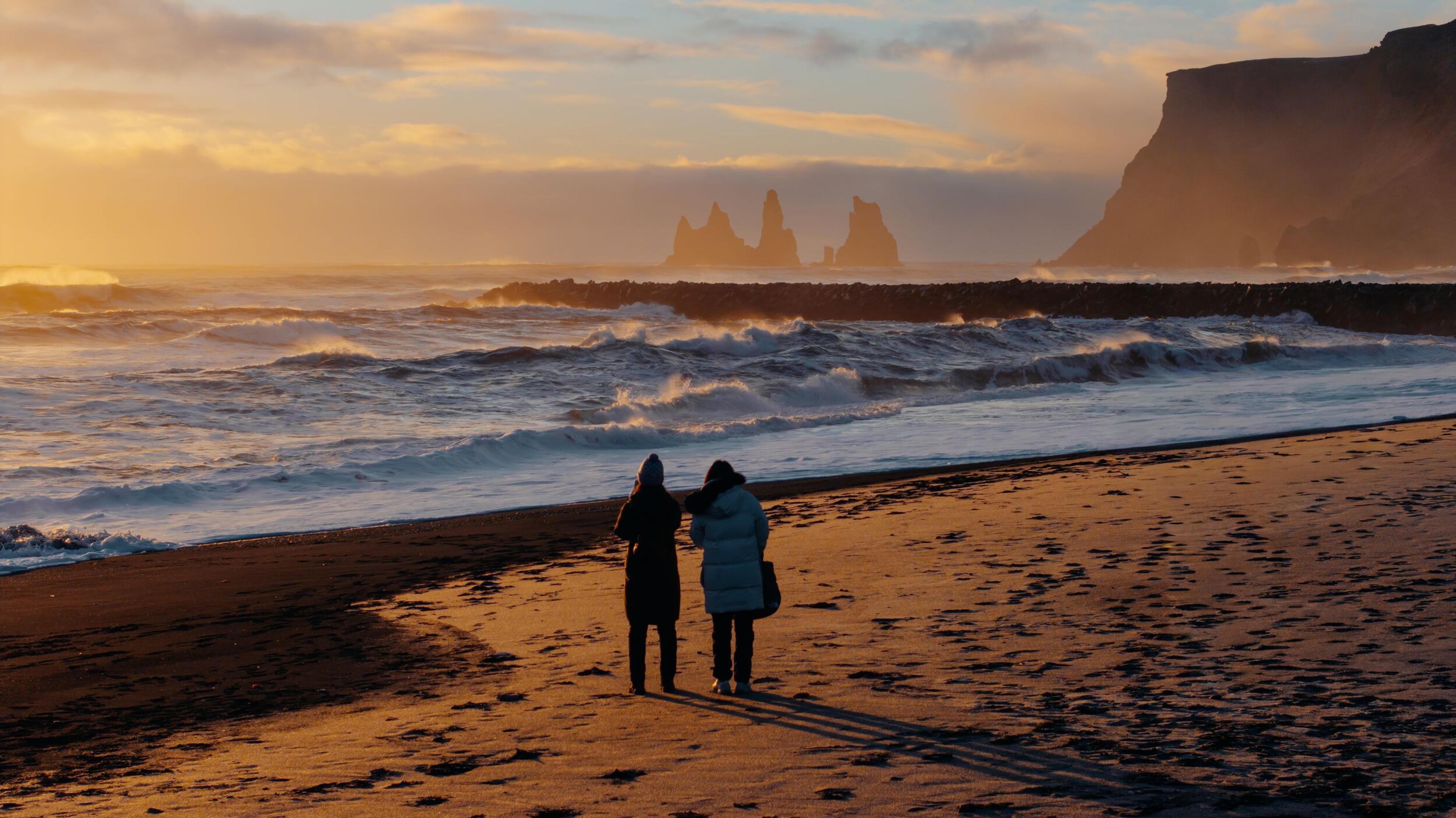 Two people stood on a black sand beach looking out at the sun setting far out at sea