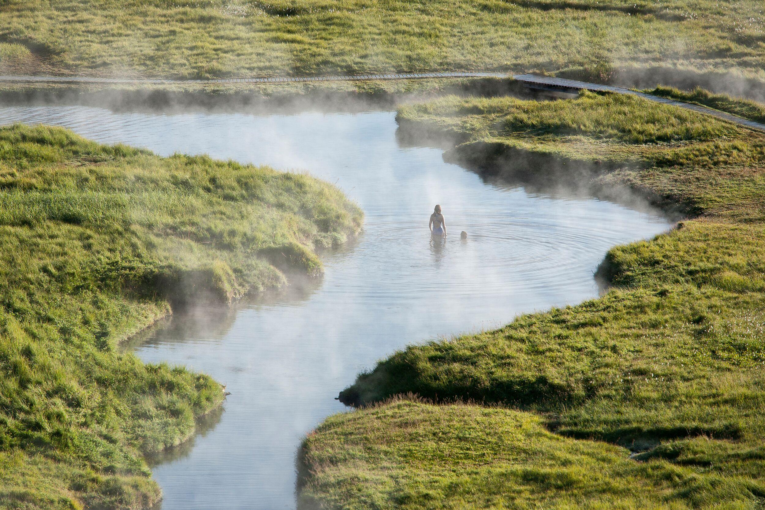 A hiker enjoying a dip in a steaming hot spring surrounded by lush greenery in Landmannalaugar, Iceland.