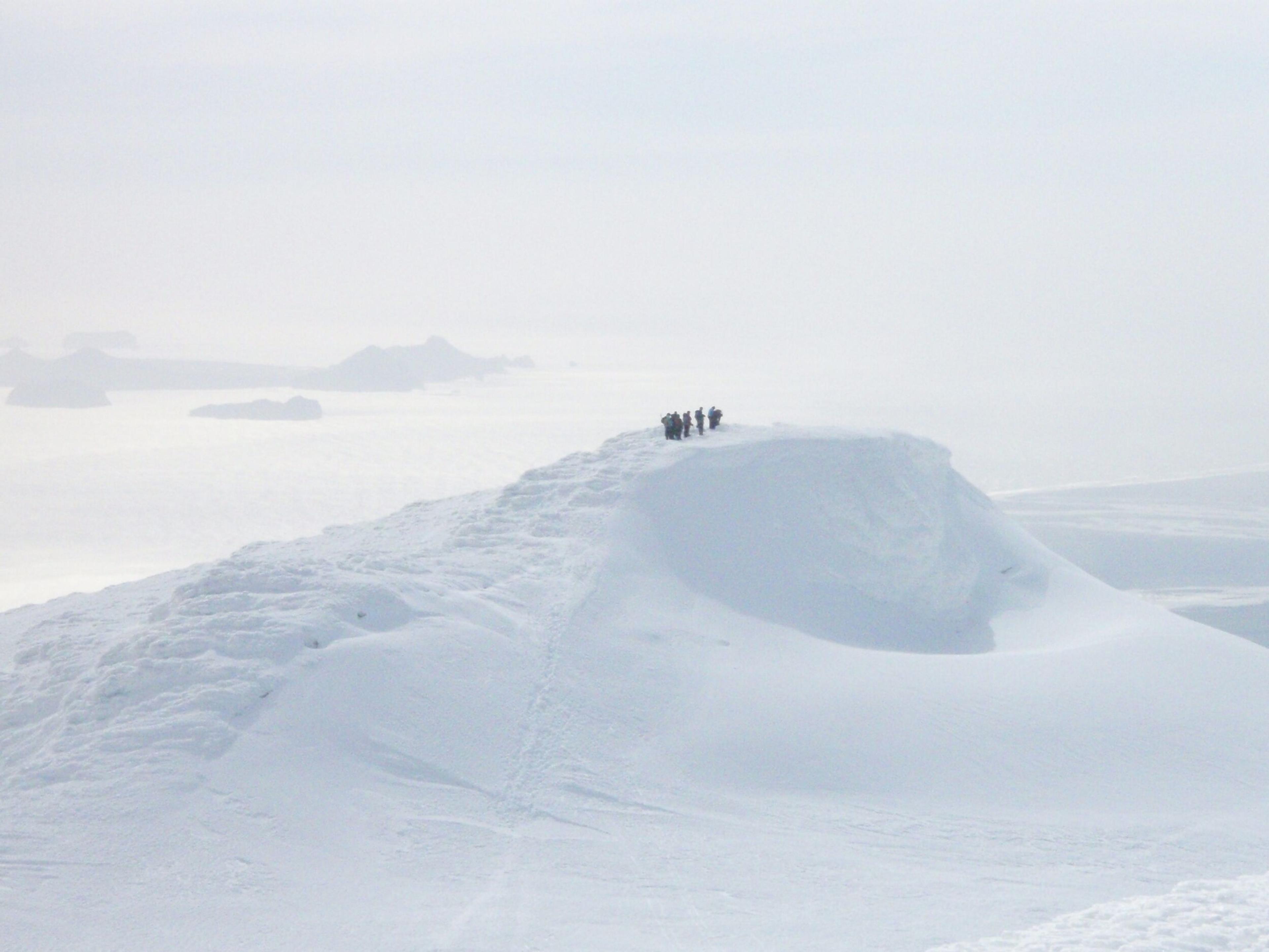 A distant view of hikers standing atop a snow-covered peak on Eyjafjallajökull, surrounded by a misty, ethereal landscape.