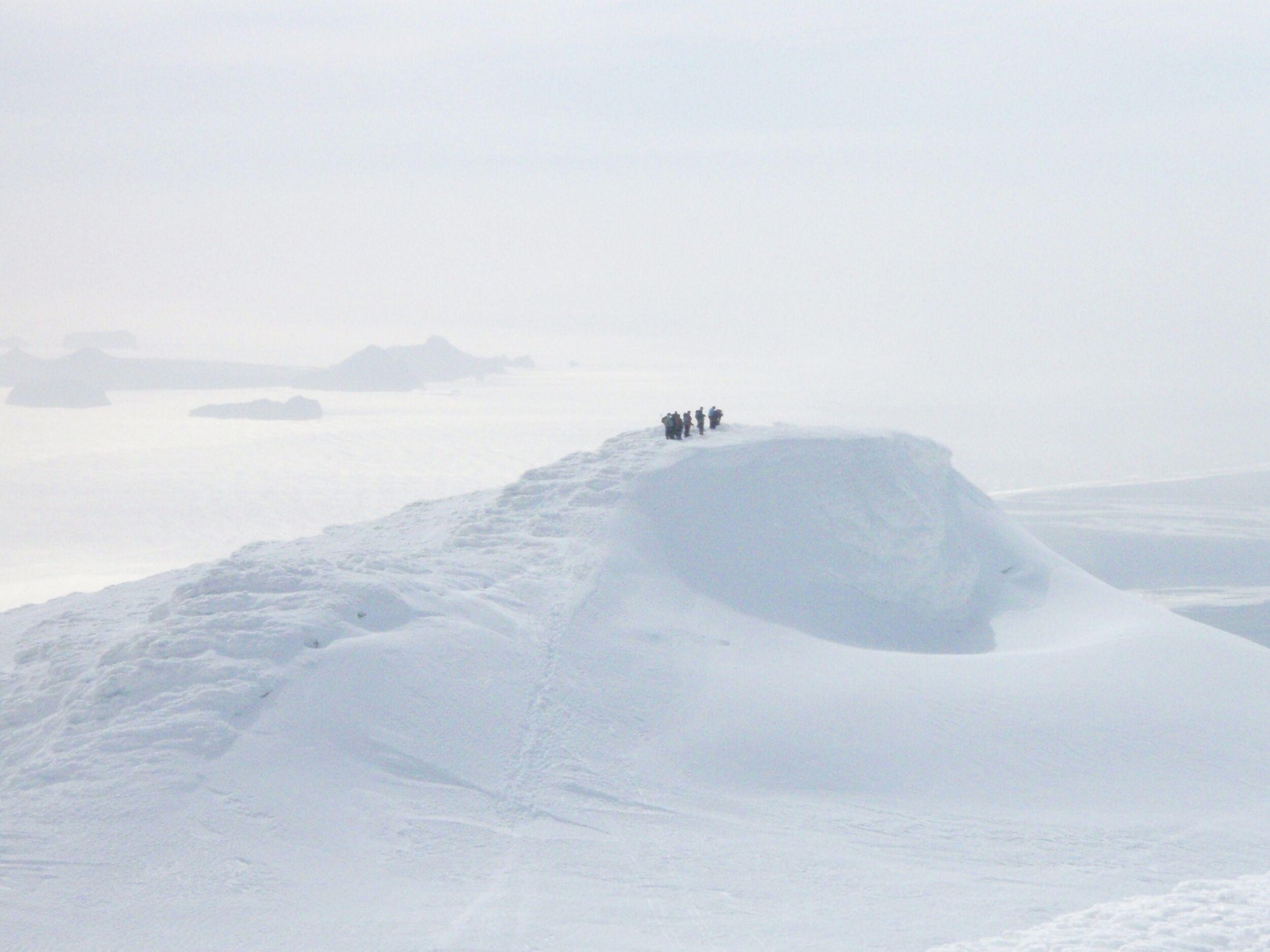A distant view of hikers standing atop a snow-covered peak on Eyjafjallajökull, surrounded by a misty, ethereal landscape.
