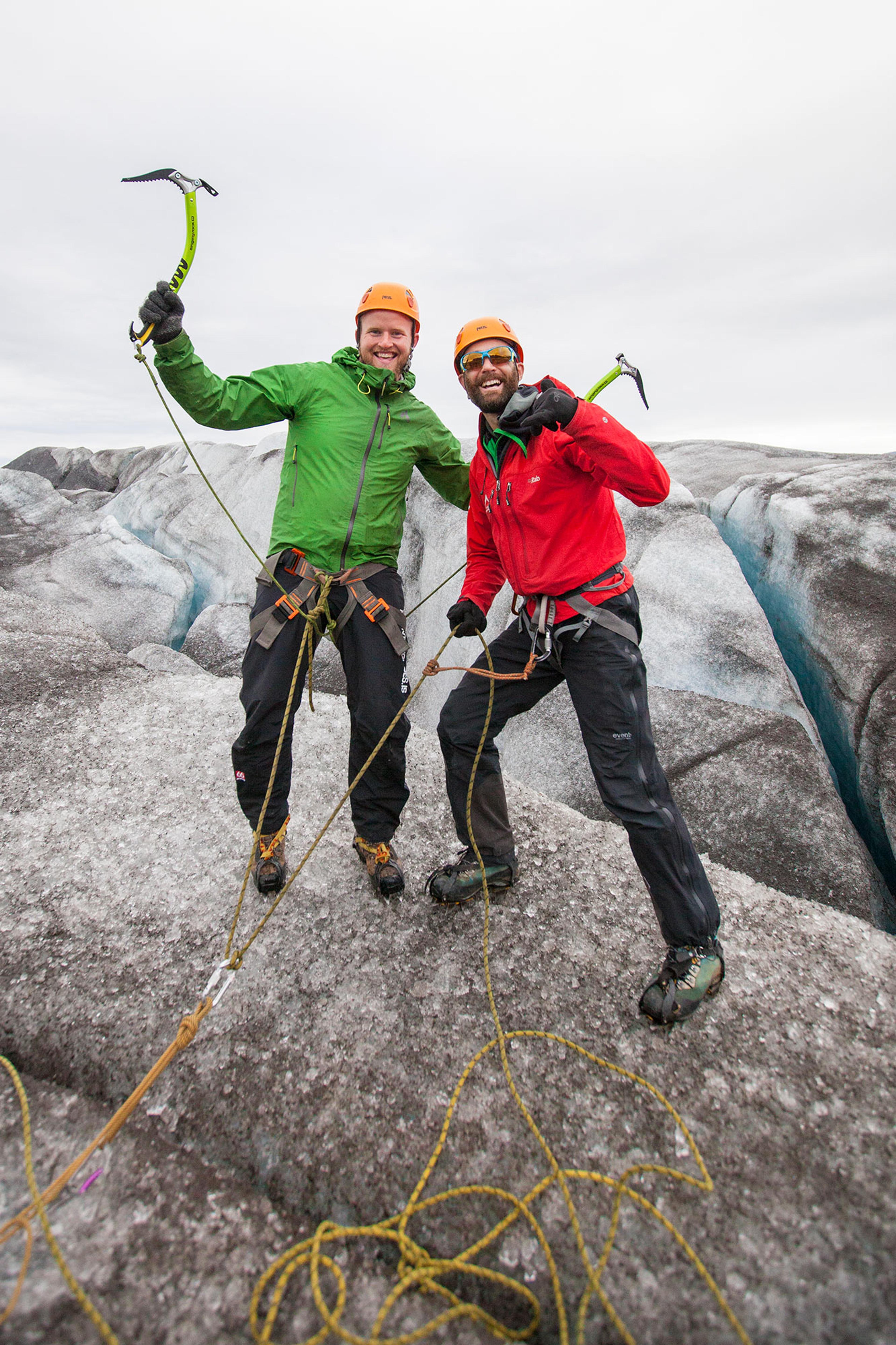 A climber in an orange helmet and jacket grins while scaling a glacier wall, using her ice axes to grip the surface.