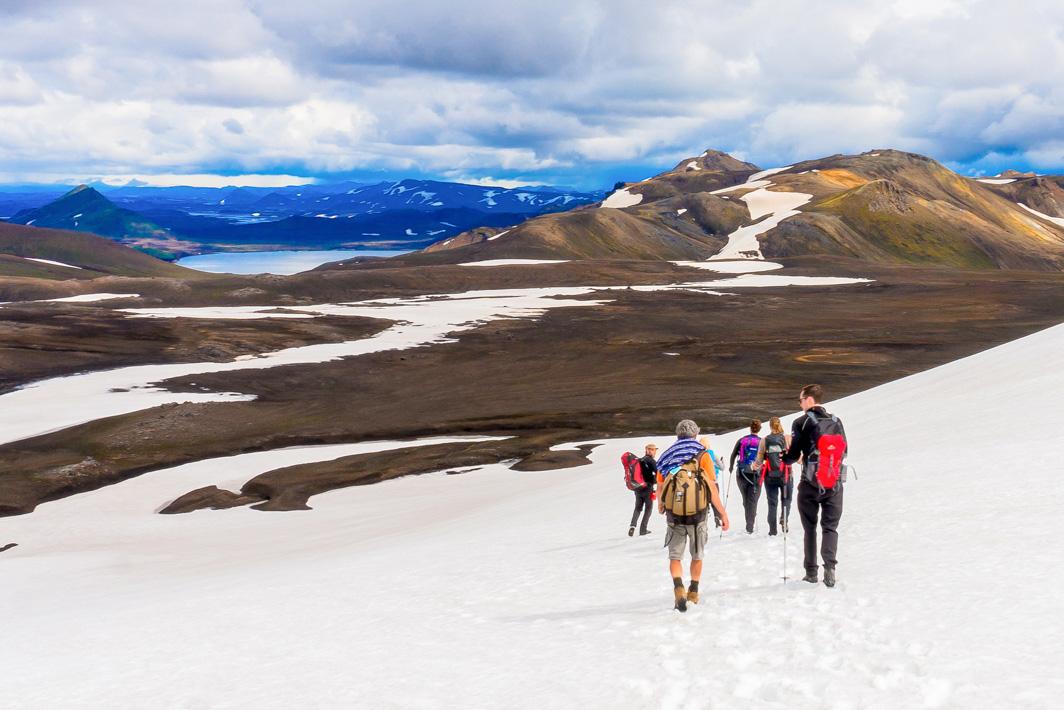 A group of hikers with backpacks traverses a snow-covered trail with patches of exposed ground, with mountainous terrain in the distance under a partly cloudy sky.
