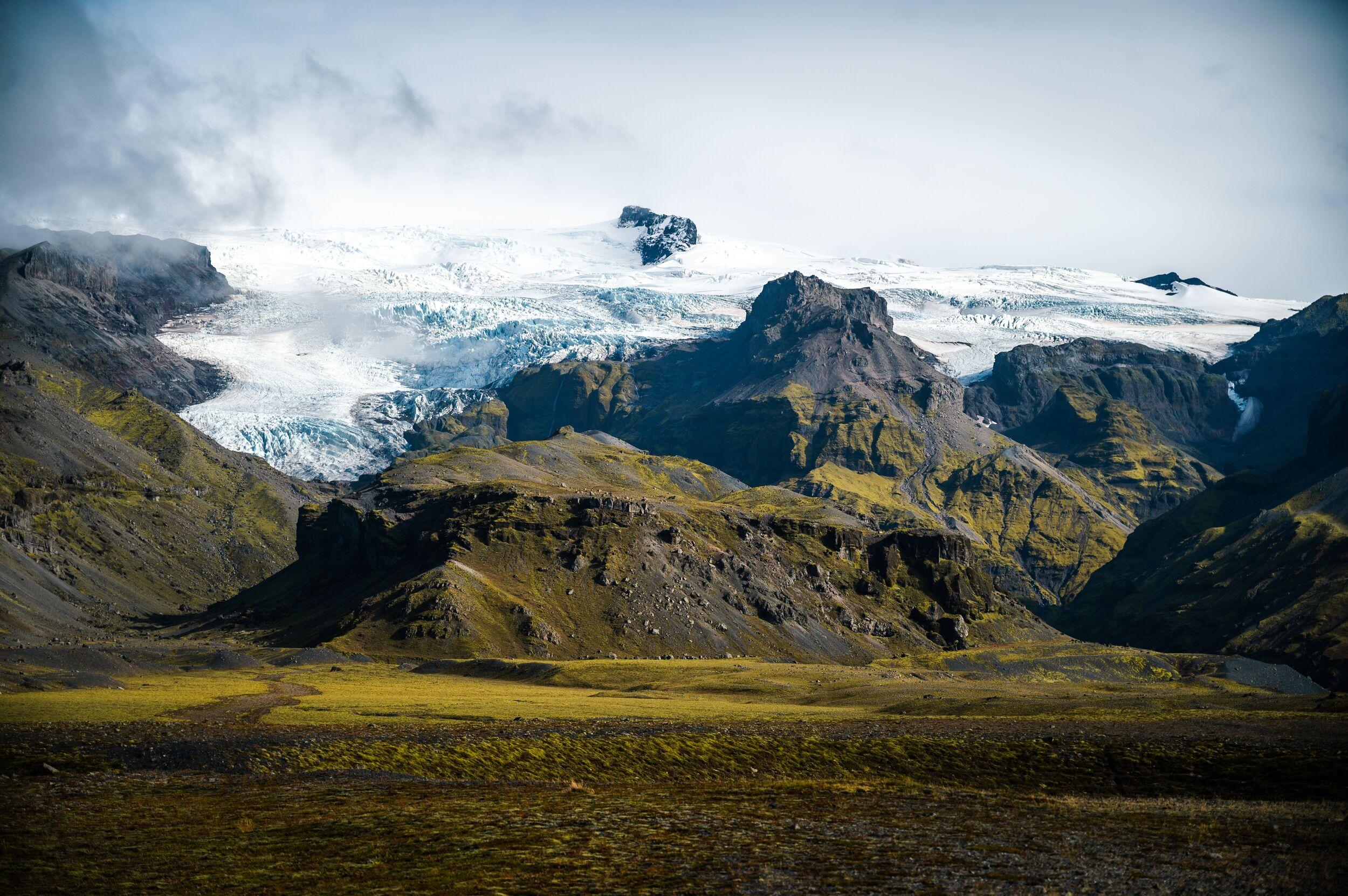 Scenic view of moss-covered volcanic mountains with a glacier cascading down between rocky peaks in Skaftafell, Iceland, under a cloudy sky.
