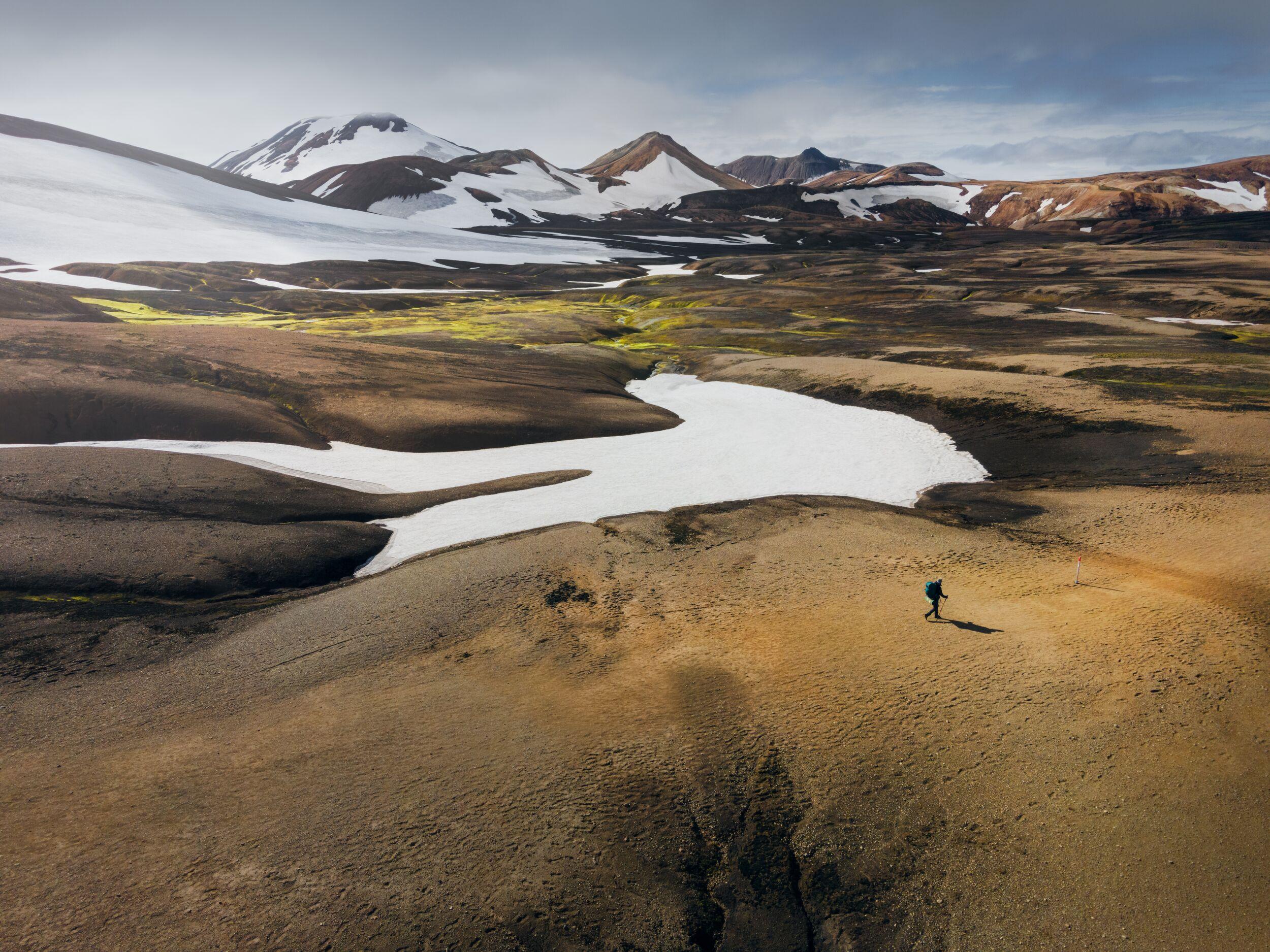 A lone hiker walks across a barren, volcanic landscape in the Icelandic Highlands, with patches of snow and distant mountains creating a striking contrast of colors and textures. The vast, open terrain emphasizes the remote and rugged nature of the area.