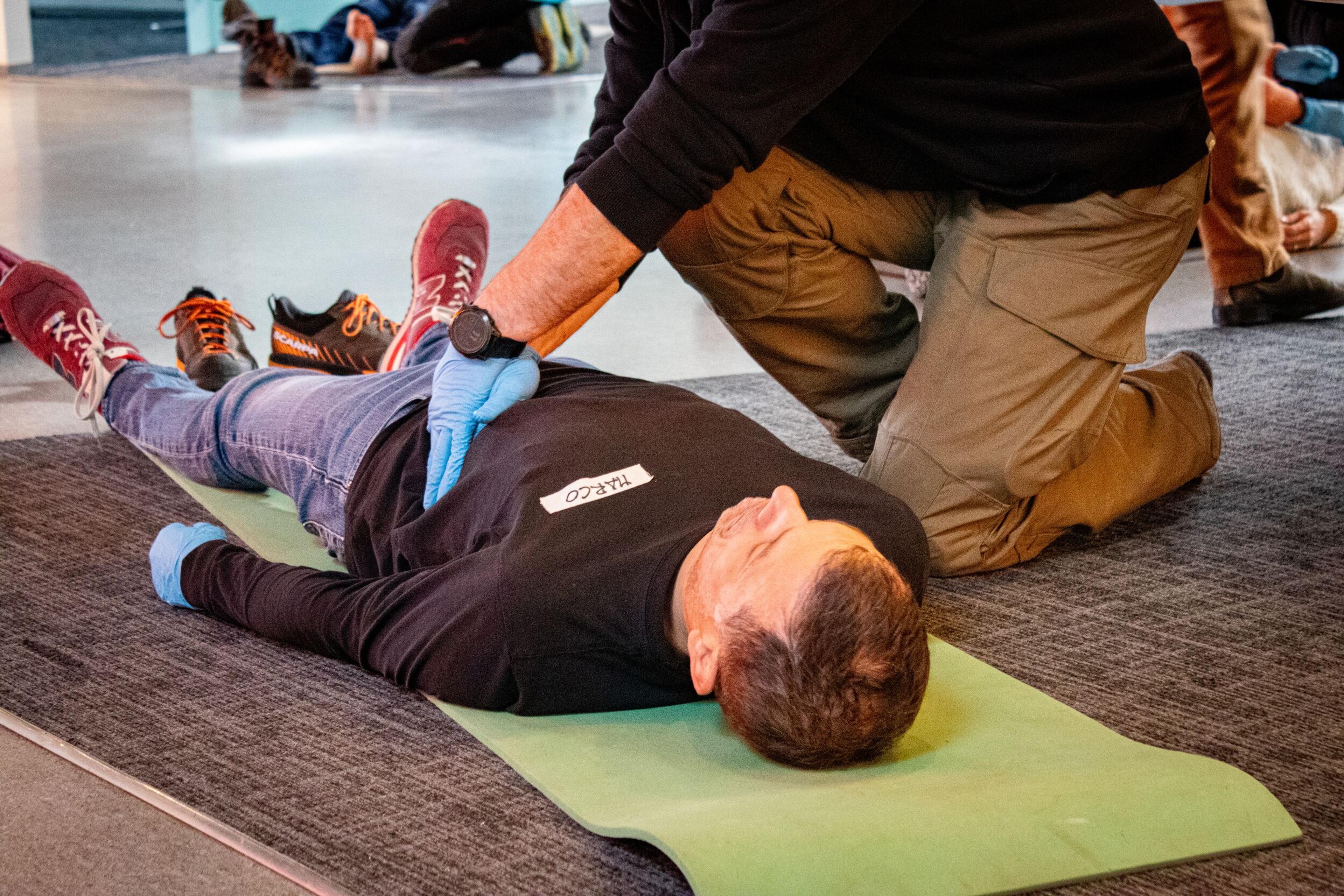 A participant in a Wilderness First Responder course practices CPR on a man lying on a mat indoors, focusing on proper hand positioning.