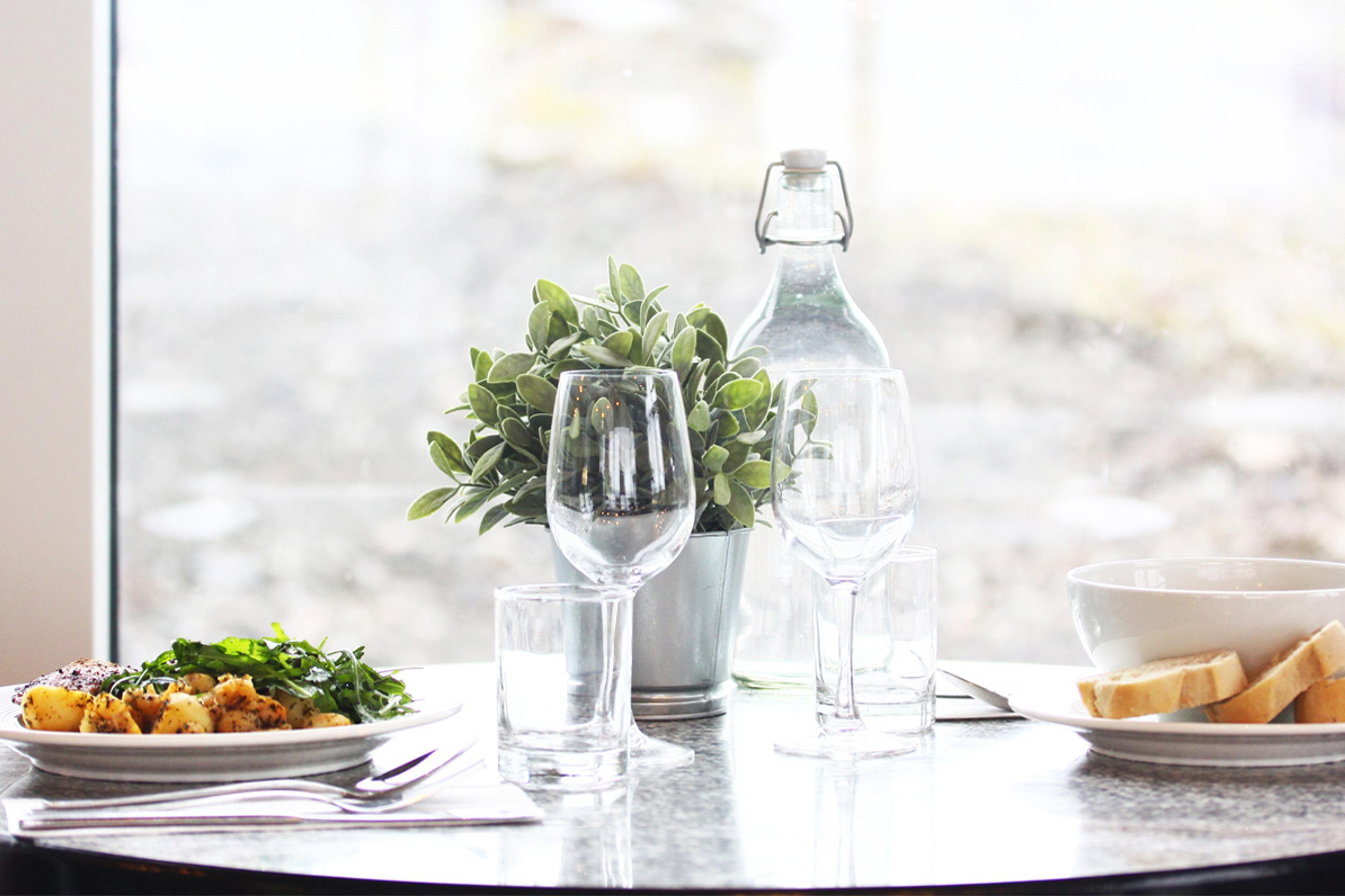 A brightly lit table with glasses, water and plates with food in front of a window.
