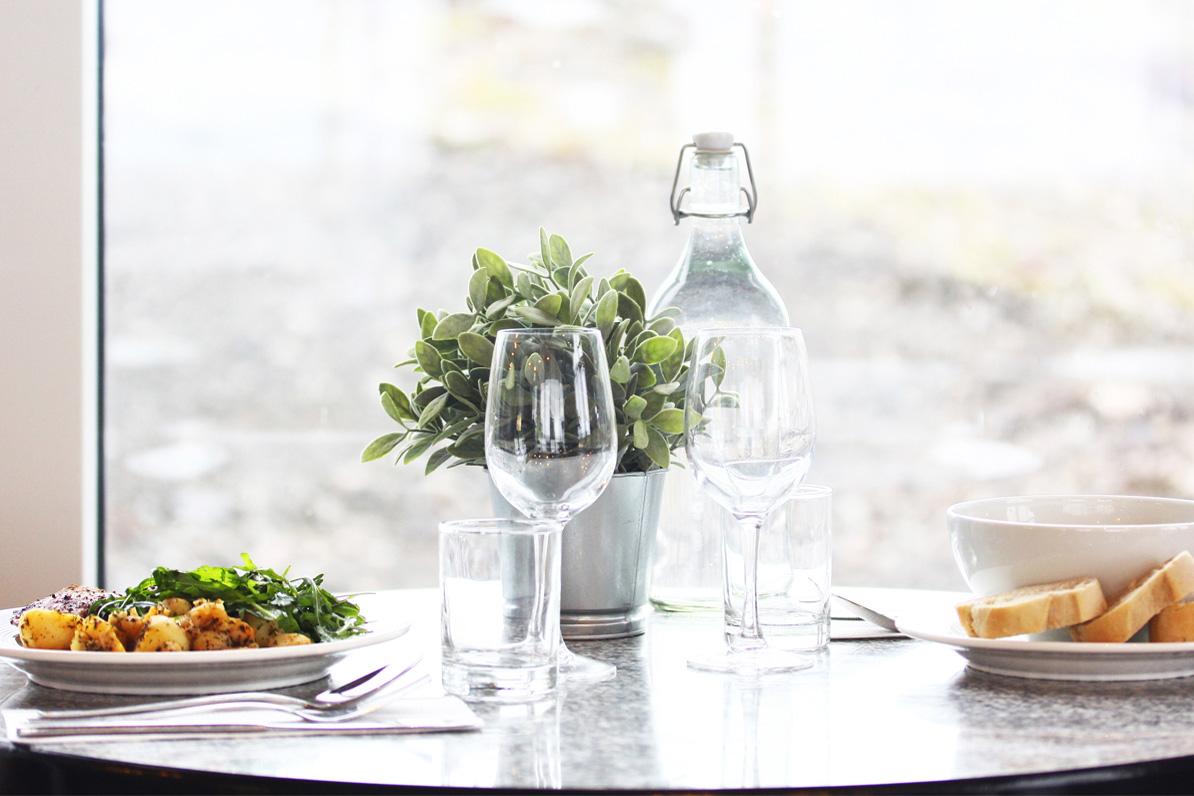 A brightly lit table with glasses, water and plates with food in front of a window.
