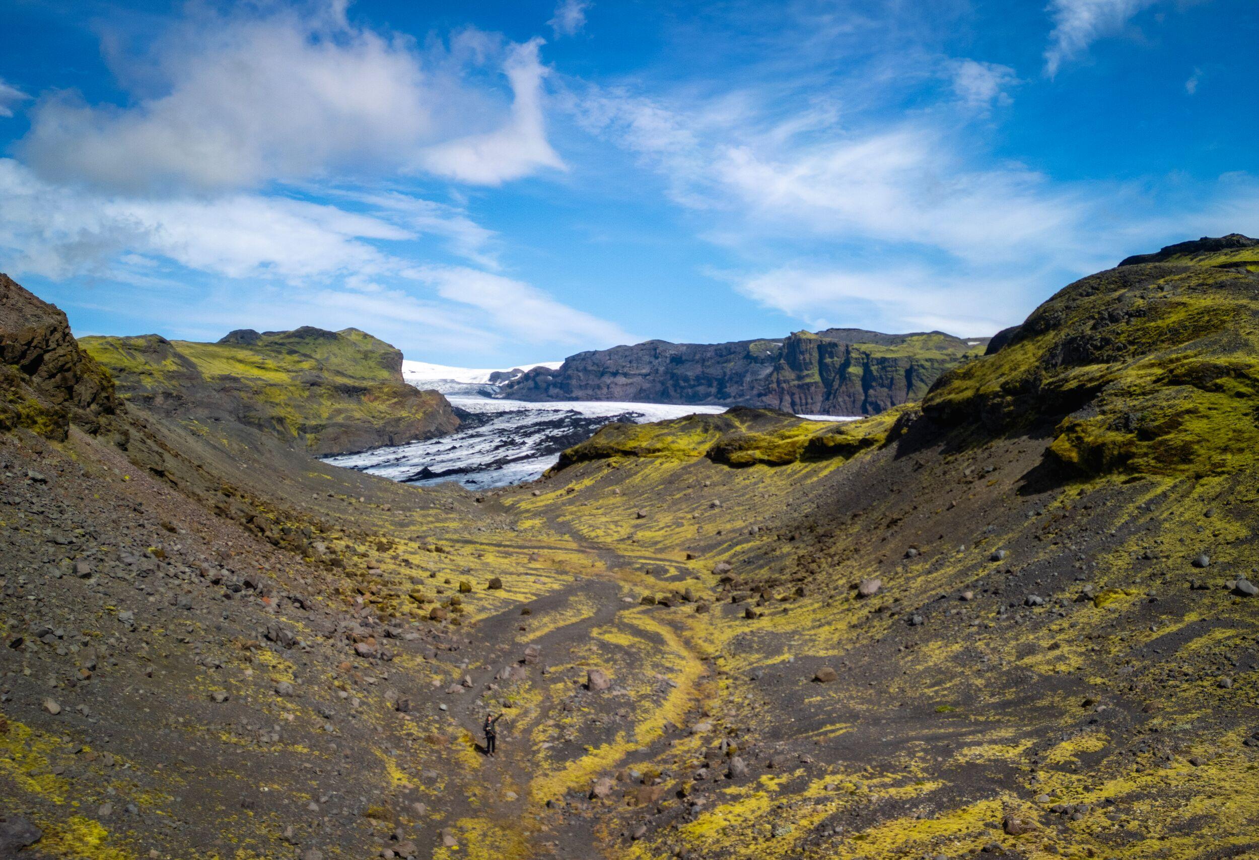 A winding trail cuts through moss-covered volcanic hills leading toward a distant glacier tongue framed by dark cliffs under a blue sky.