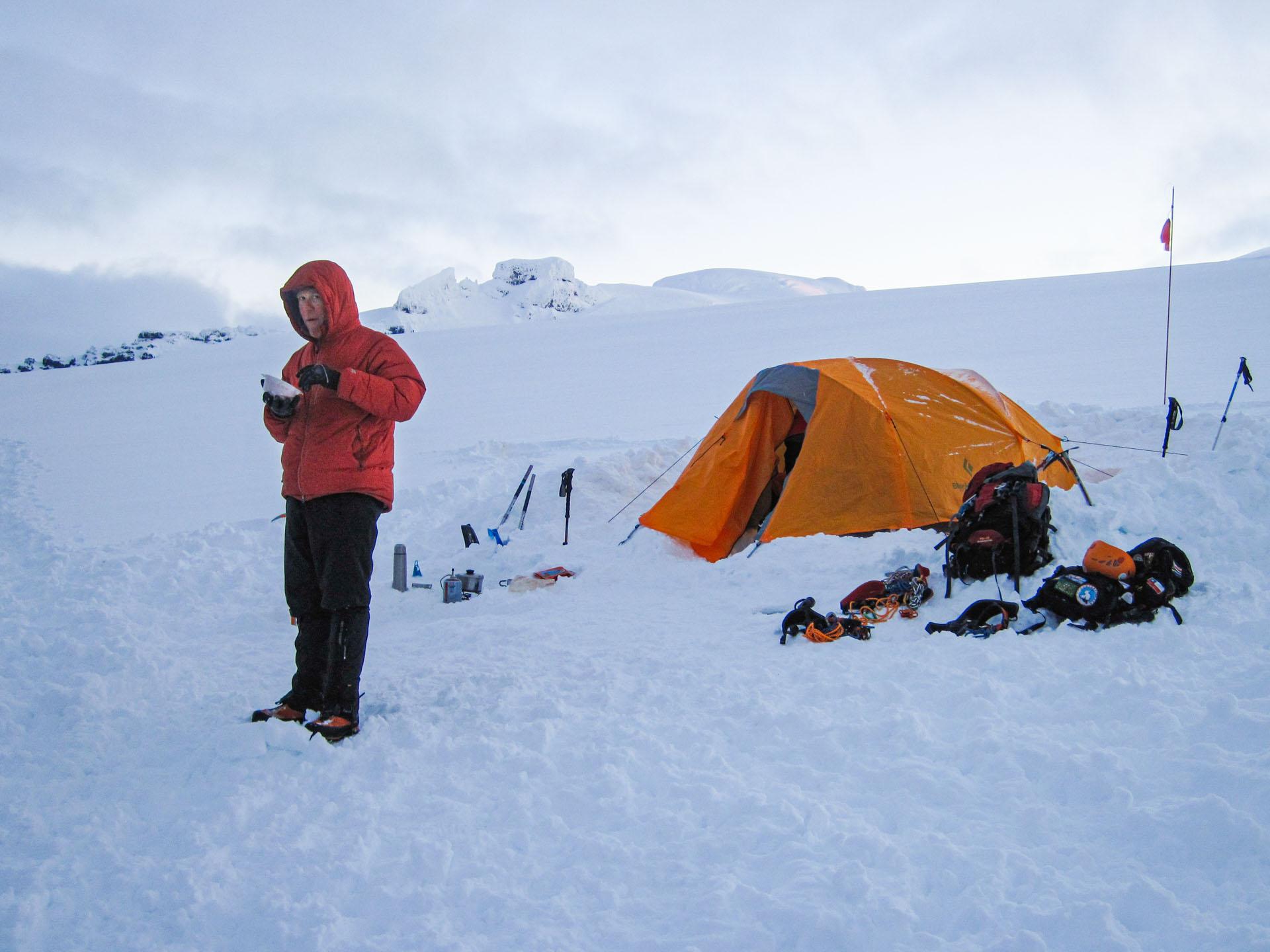 man eating on the cold snowy mountain with a orange tent behind