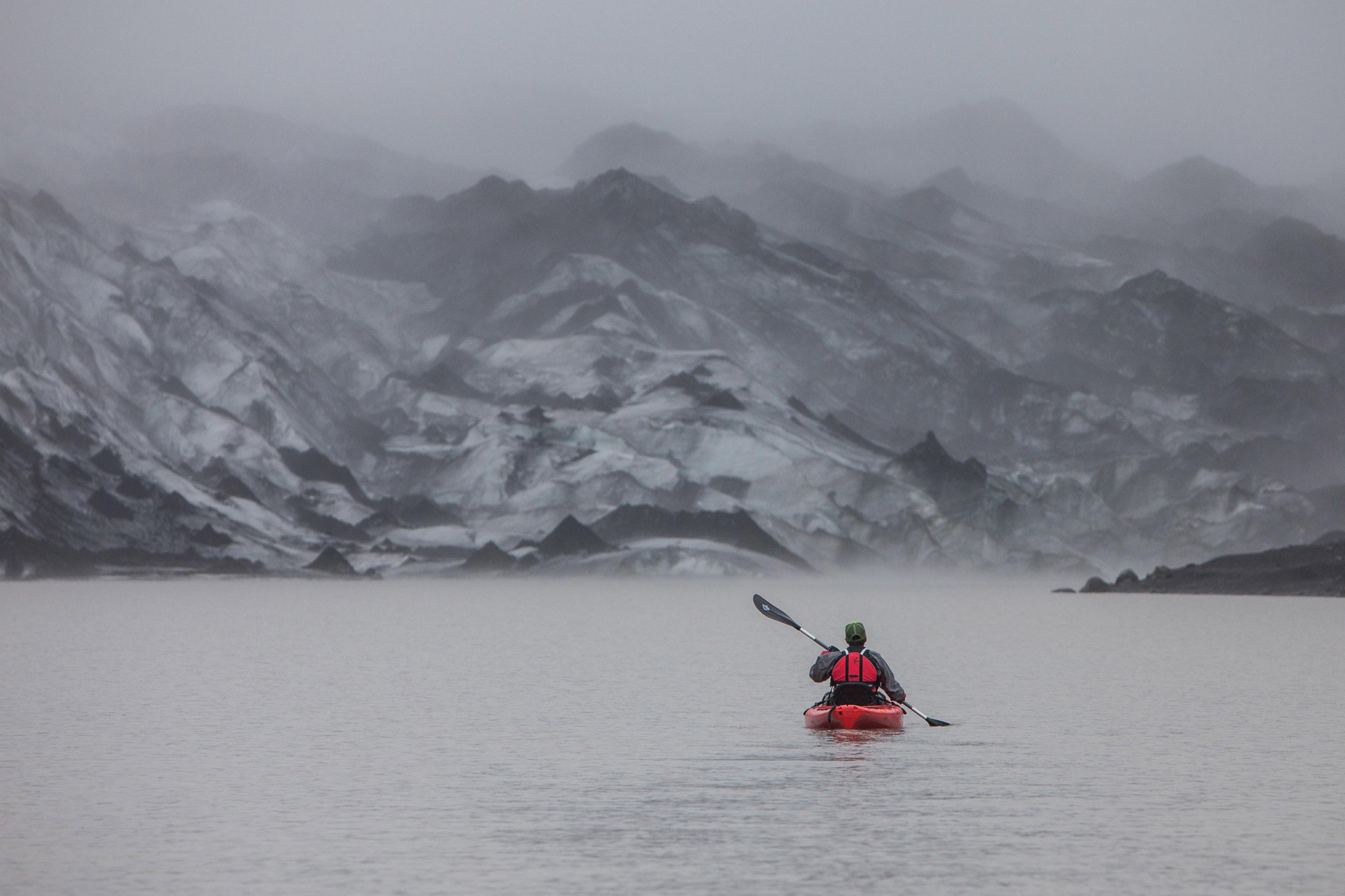 A person in a red kayak paddling on a glacier lagoon toward a glacier that is partially covered in fog.