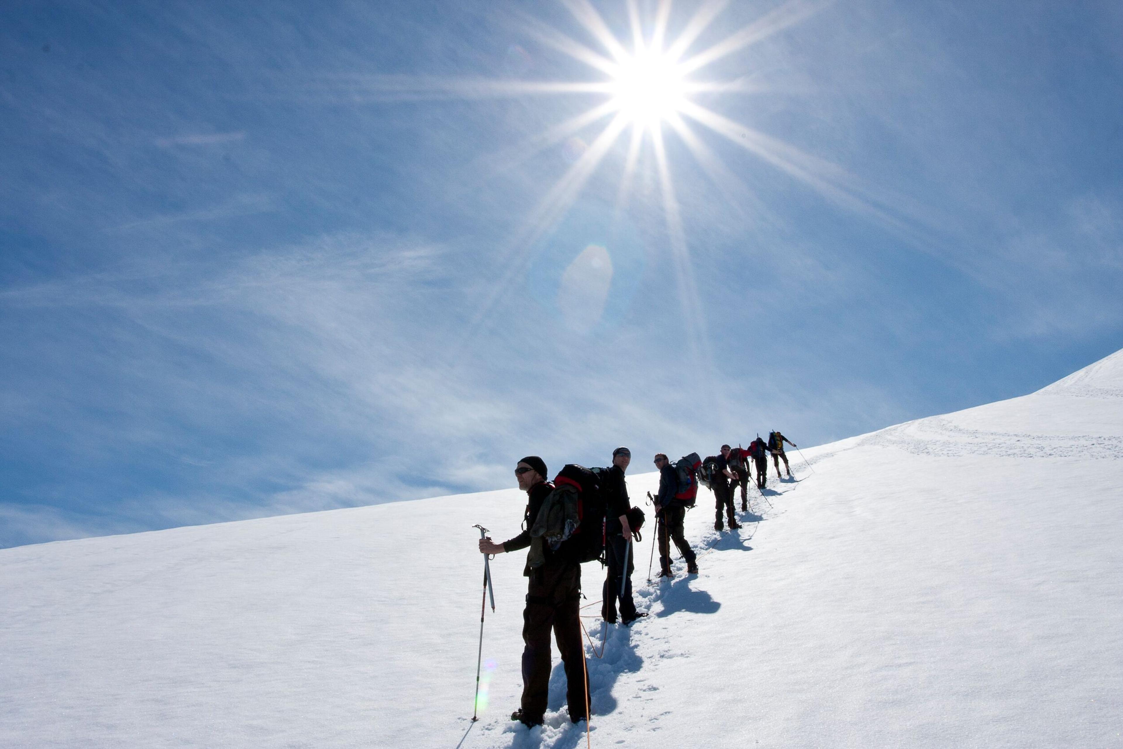 A line of hikers ascends a snowy slope on Eyjafjallajökull under a bright sun and clear blue sky, each using trekking poles for stability.