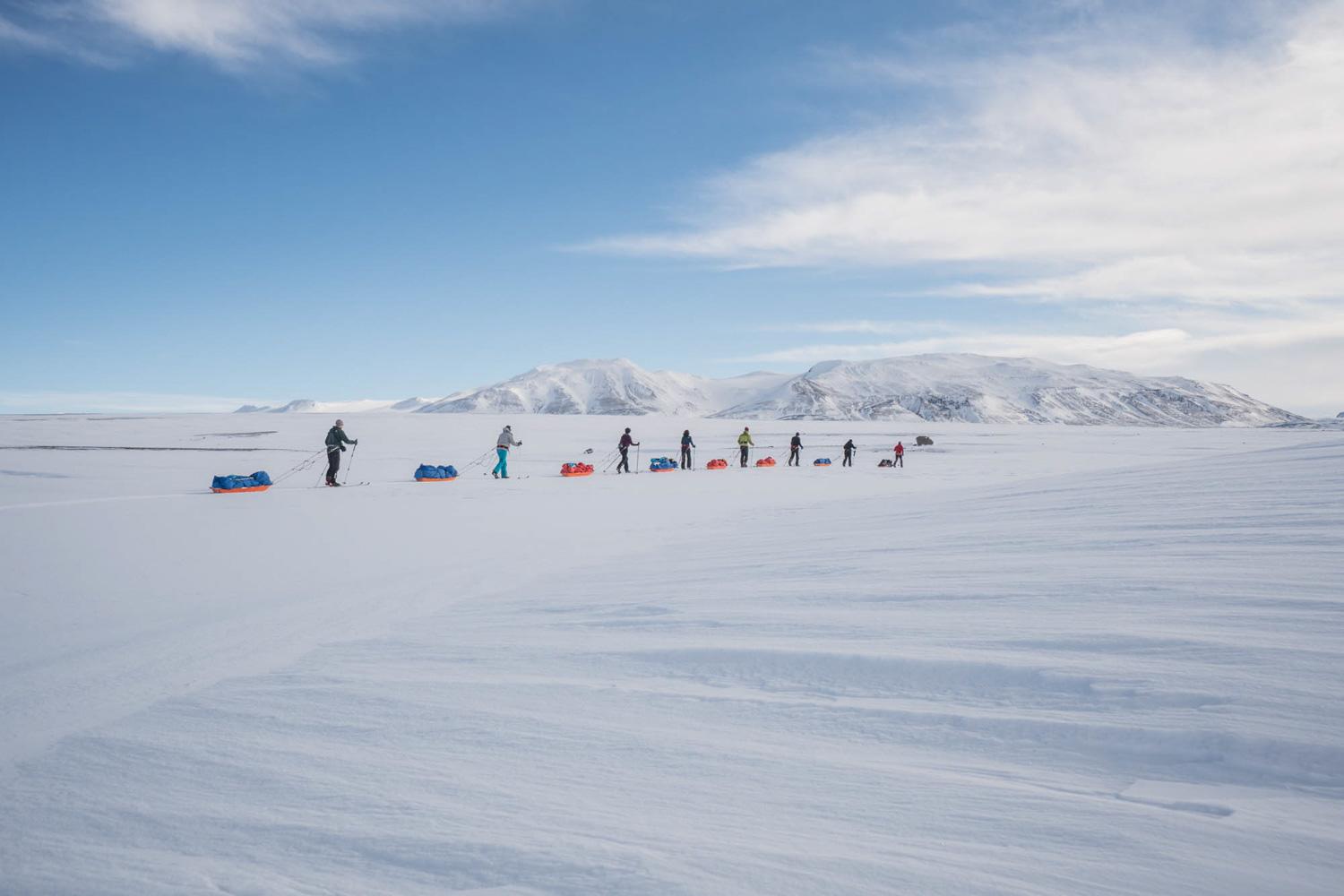 A line of trekkers in winter gear traverses a snowy landscape with a backdrop of mountainous terrain.