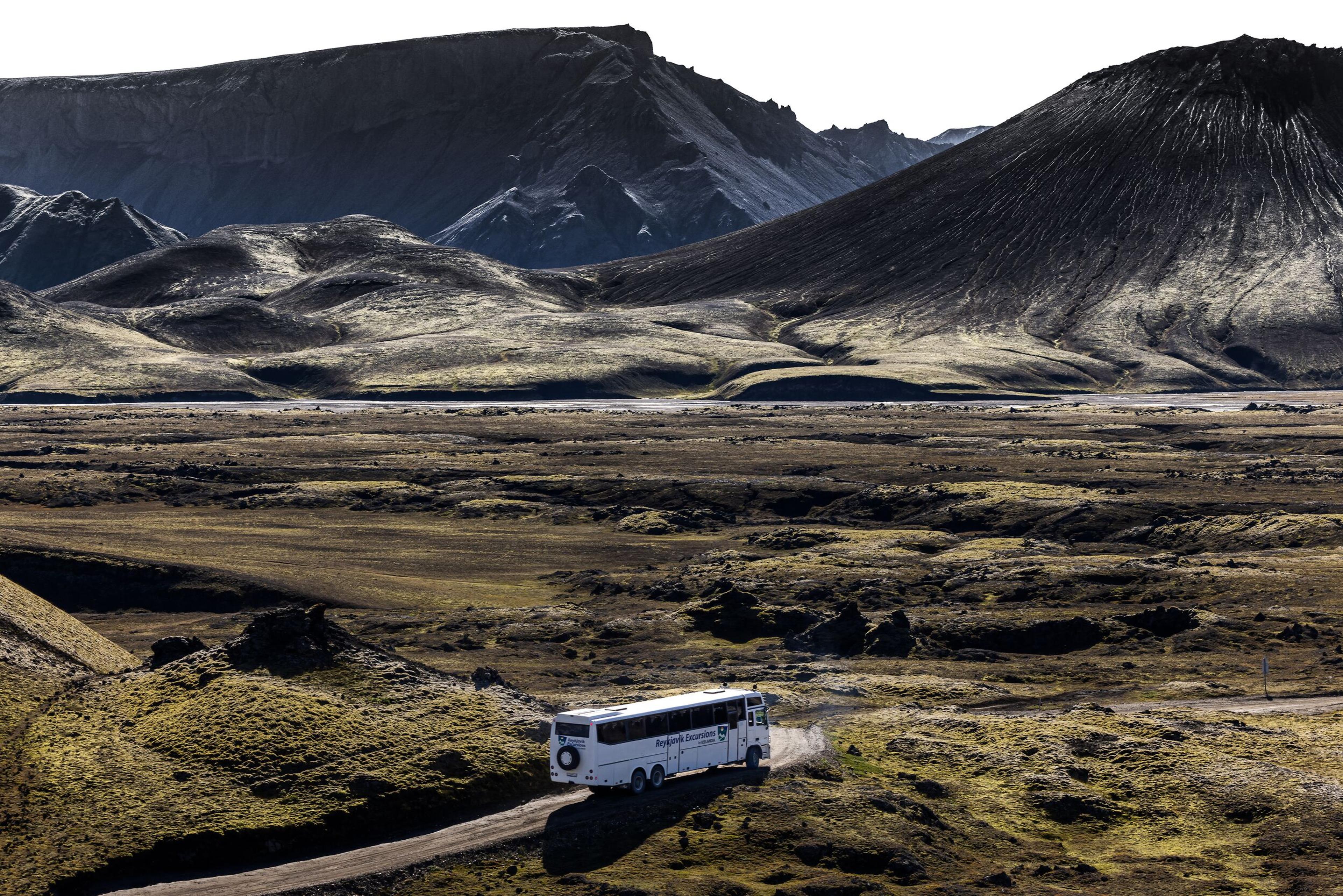 A white tour bus drives along a dirt road through the vast, rugged landscape of the Icelandic Highlands, surrounded by volcanic hills and dark, craggy mountains.