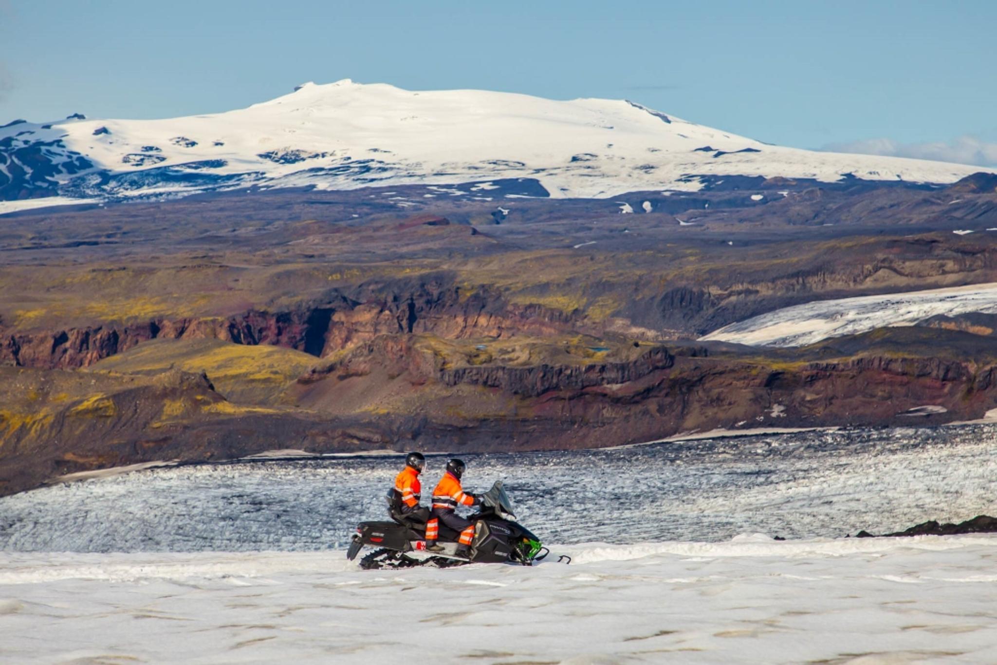 Two people on a snowmobile riding on a vast snowy terrain with an ice-capped mountain looming in the beackground
