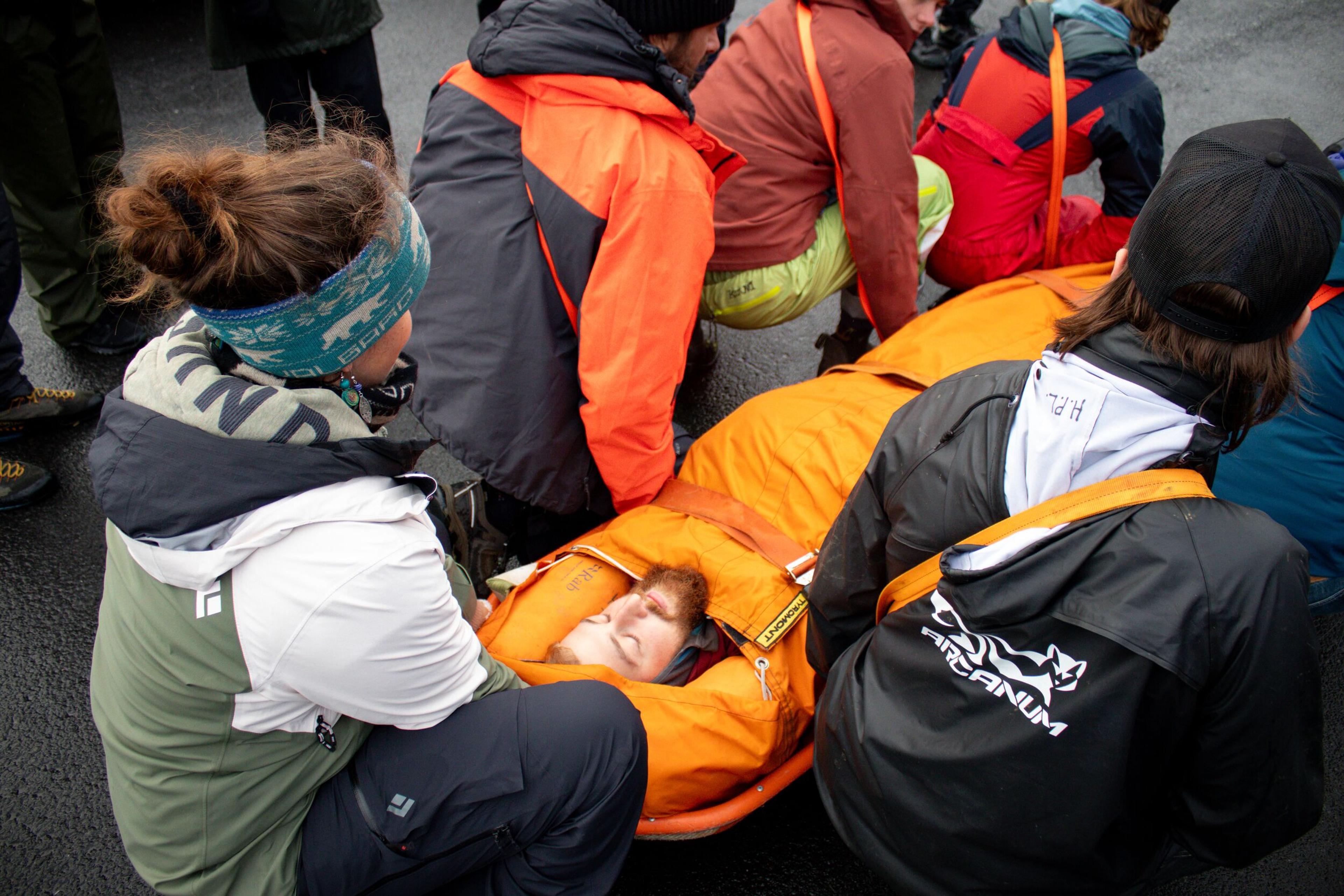 A group of Wilderness First Responder course participants prepare a person wrapped in an orange stretcher for transport, simulating an emergency rescue scenario.