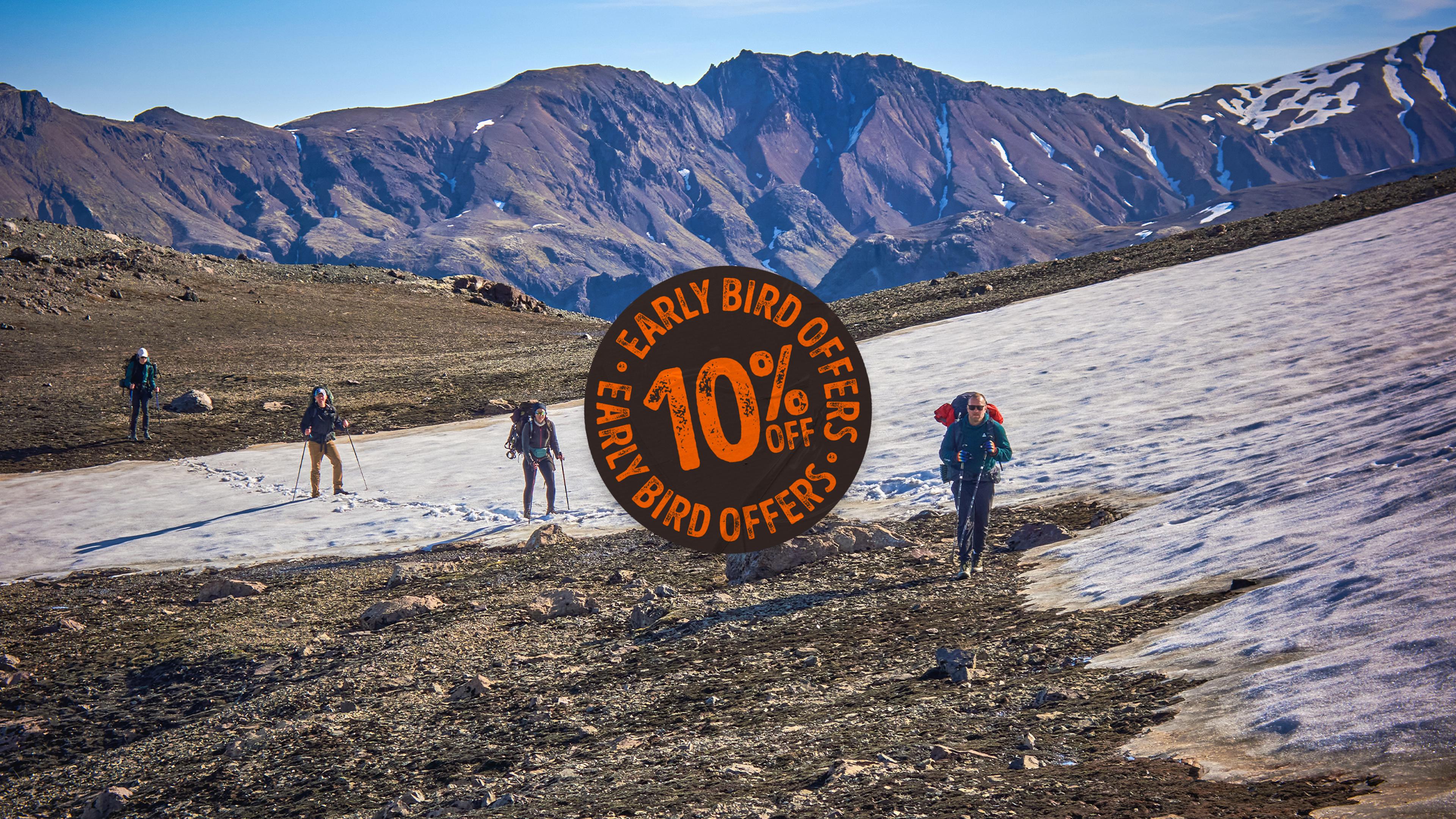 This image shows a person wearing colorful pants, a backpack, and a bright orange beanie hiking across a stunning glacier. The icy blue surface of the glacier contrasts with the dark mountains and snow-capped peaks in the background, evoking a sense of adventure in a remote and frigid landscape
