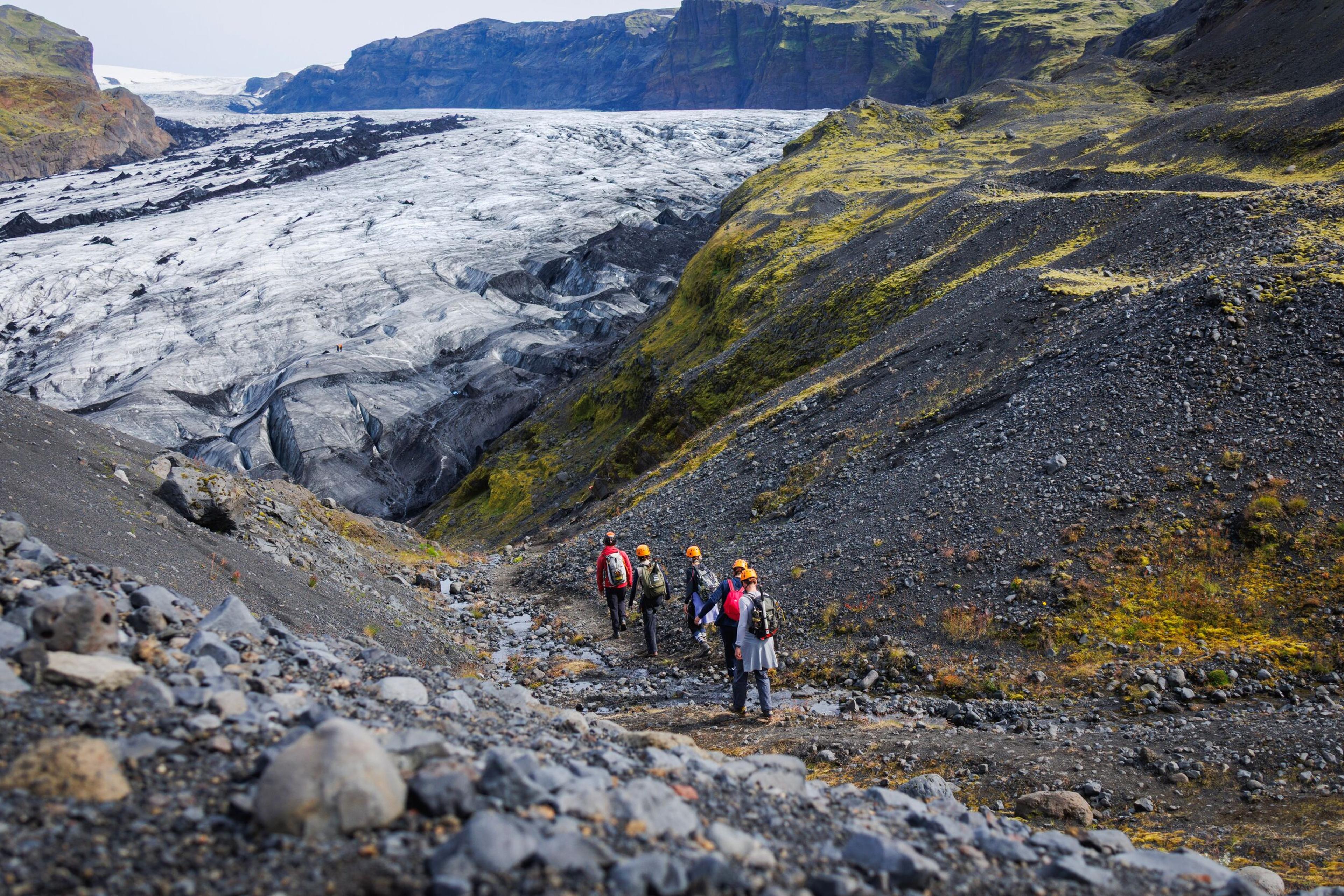 Hikers in orange helmets walk a rocky trail alongside a massive crevassed glacier, with sea cliffs visible in the distance.
