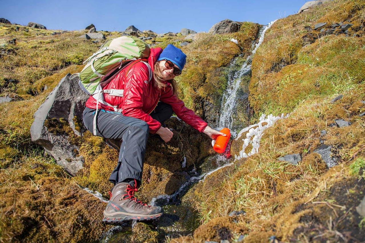 Woman in a red jacket with sunglasses getting water in a bottle from a small creek on a hiking tour in Iceland