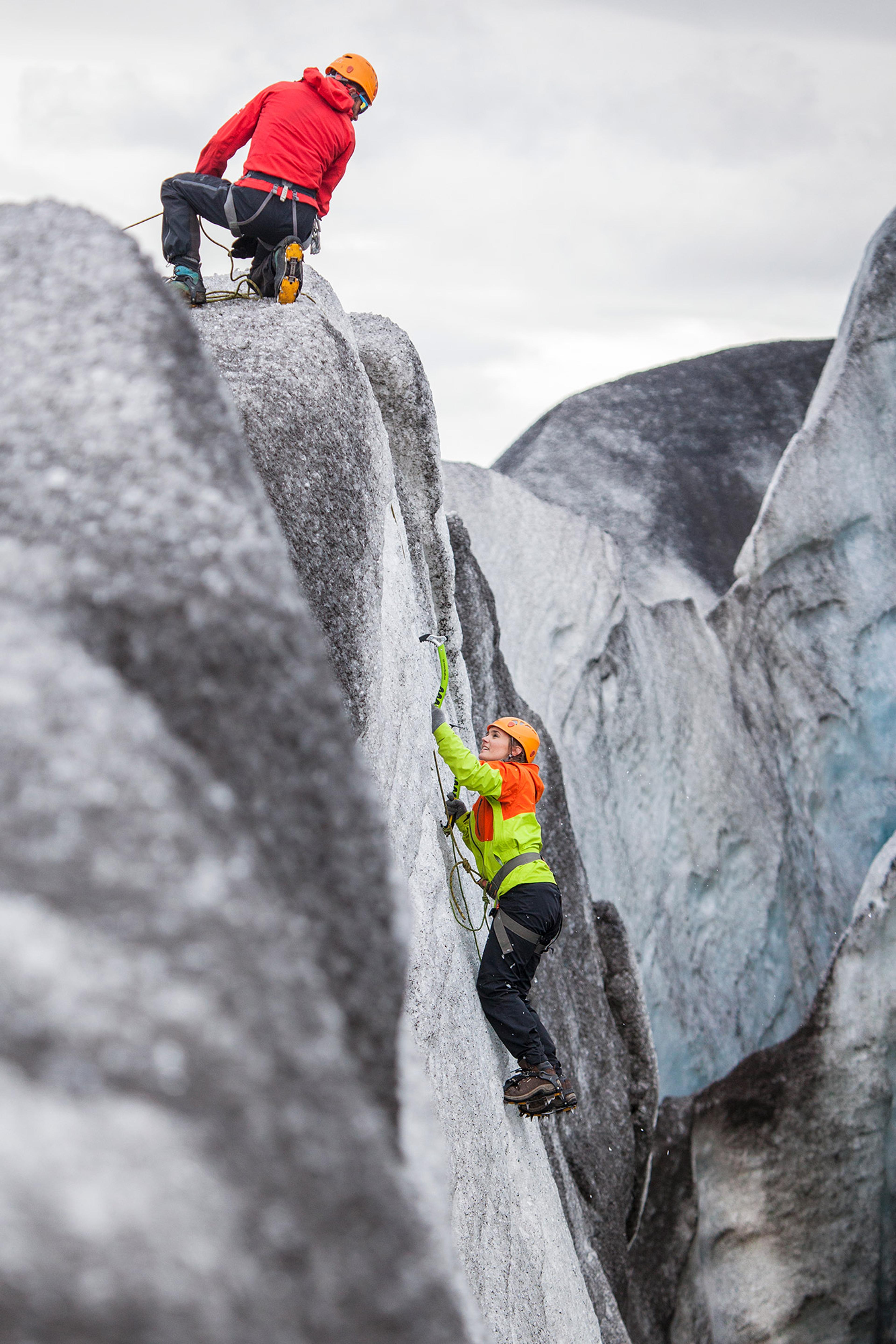 A climber in bright gear tackles a steep ice ridge, while a partner waits above on the glacier summit.