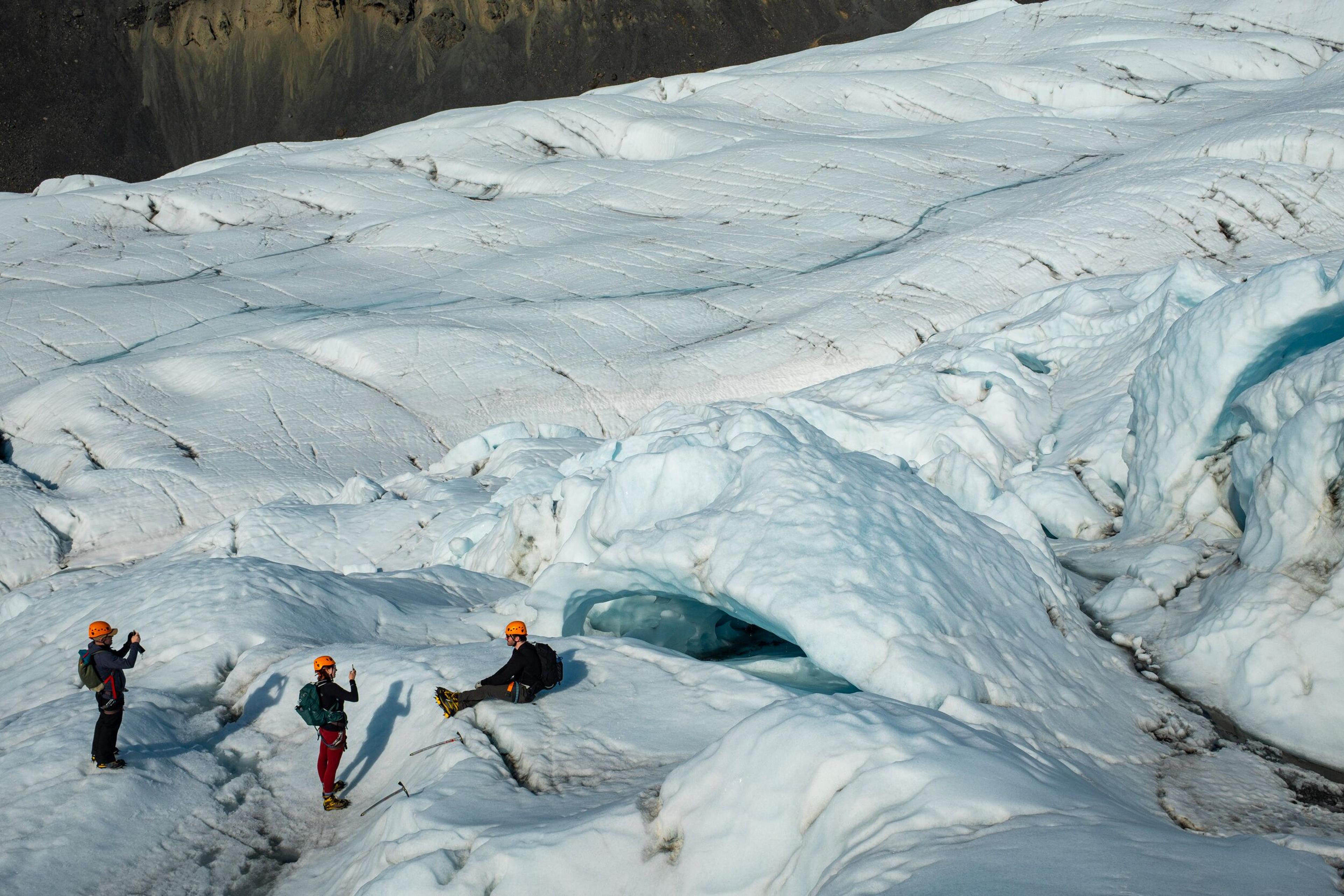 Three mountain climbers in helmets and climbing gear explore a crevasse on a vast snow-covered glacier, surrounded by rugged blue-white ice formations.