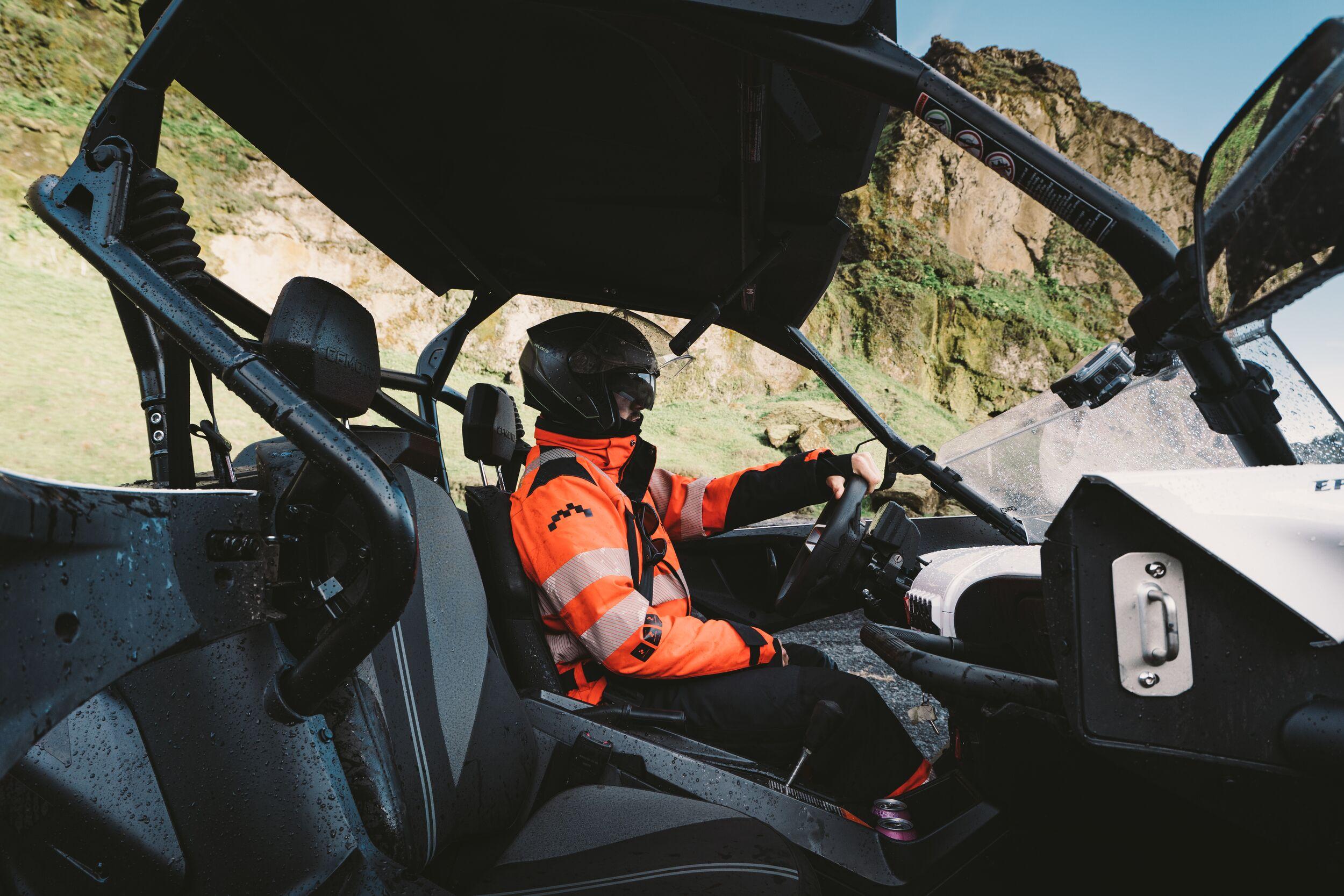 Person wearing a helmet and bright orange gear driving a buggy through rugged terrain in Iceland, with green cliffs in the background.