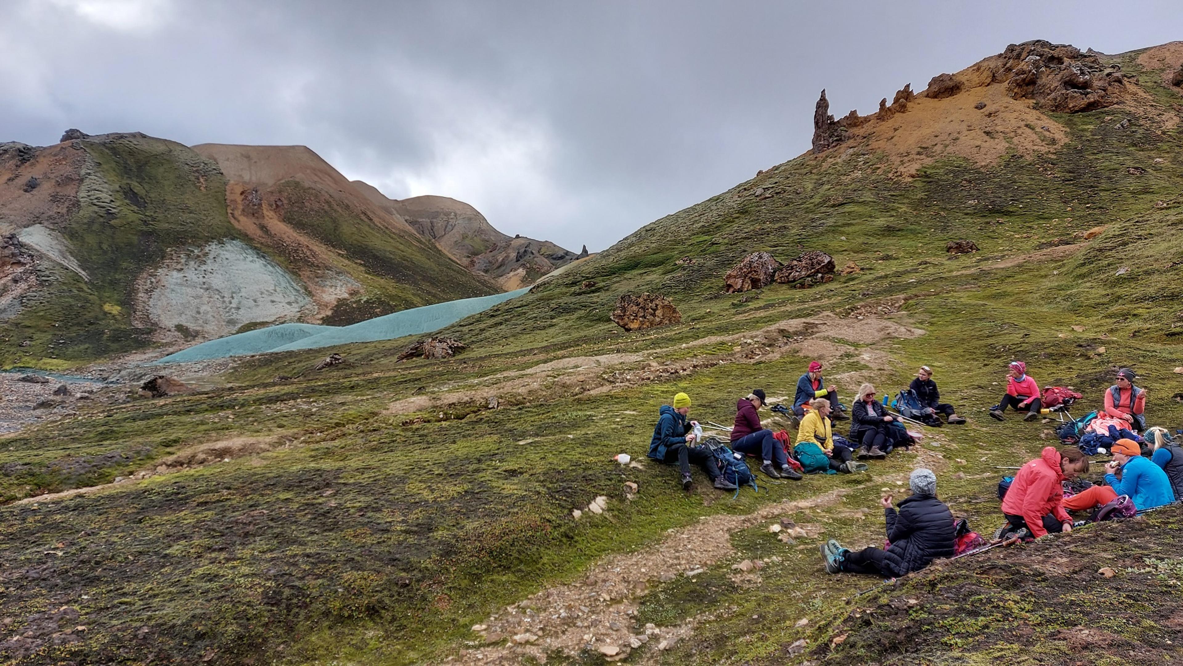 A group of hikers rests on a grassy slope surrounded by vibrant rhyolite hills and the iconic green ridges of Landmannalaugar. A line of hikers ascends the striking green hills of Landmannalaugar