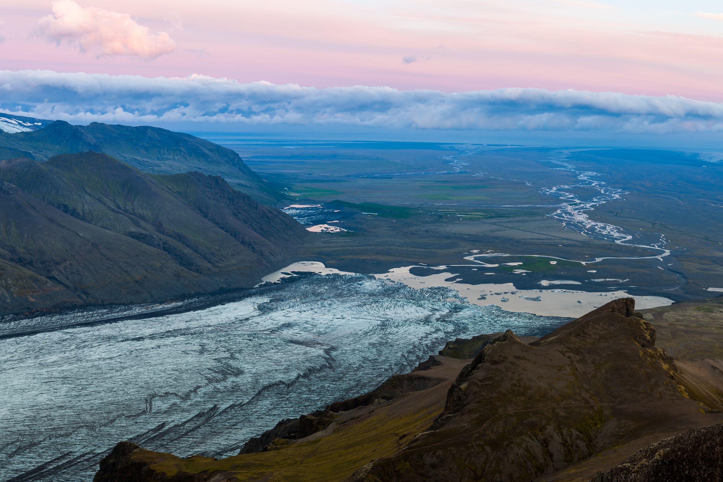 Panoramic view of a glacier winding through a wide valley beneath Kristínartindar at sunset.