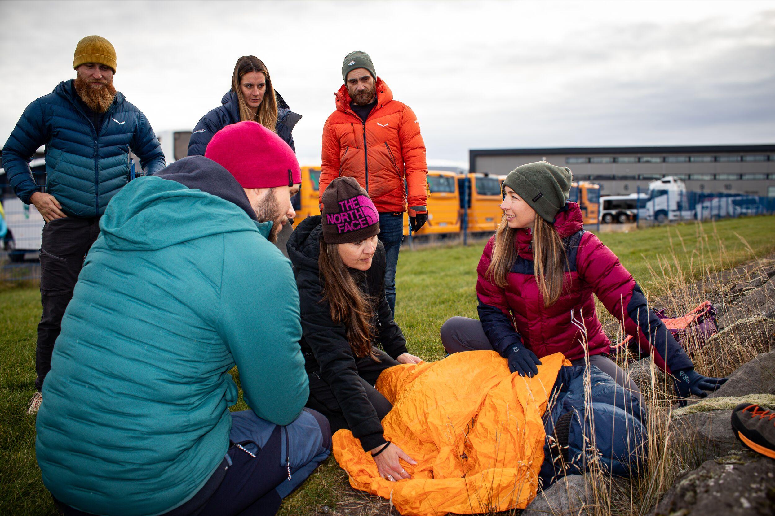 A group of Wilderness First Responder course participants gather around a simulated patient wrapped in an orange blanket, practicing outdoor emergency response techniques in a grassy area.