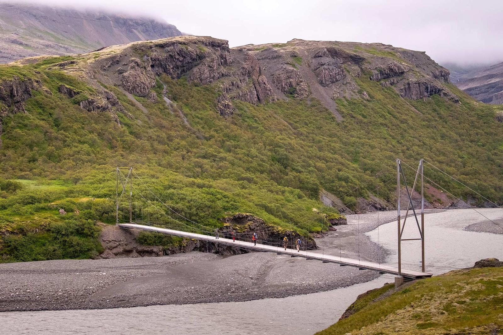 People crossing a hanging bridge over a glacial river in Lonsoraefi area in Iceland