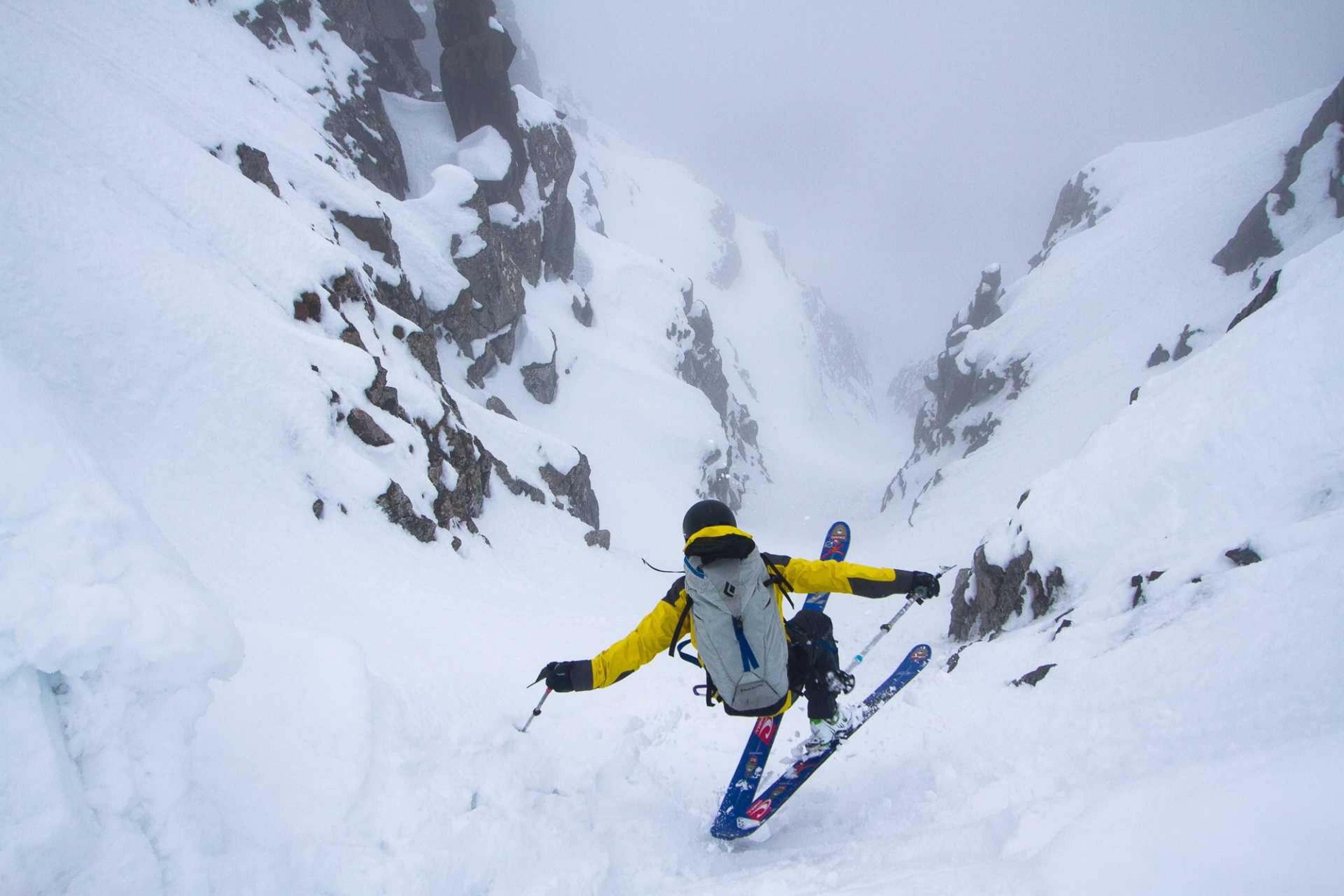 A skier descends a steep, narrow snow-filled couloir, framed by jagged rock walls.