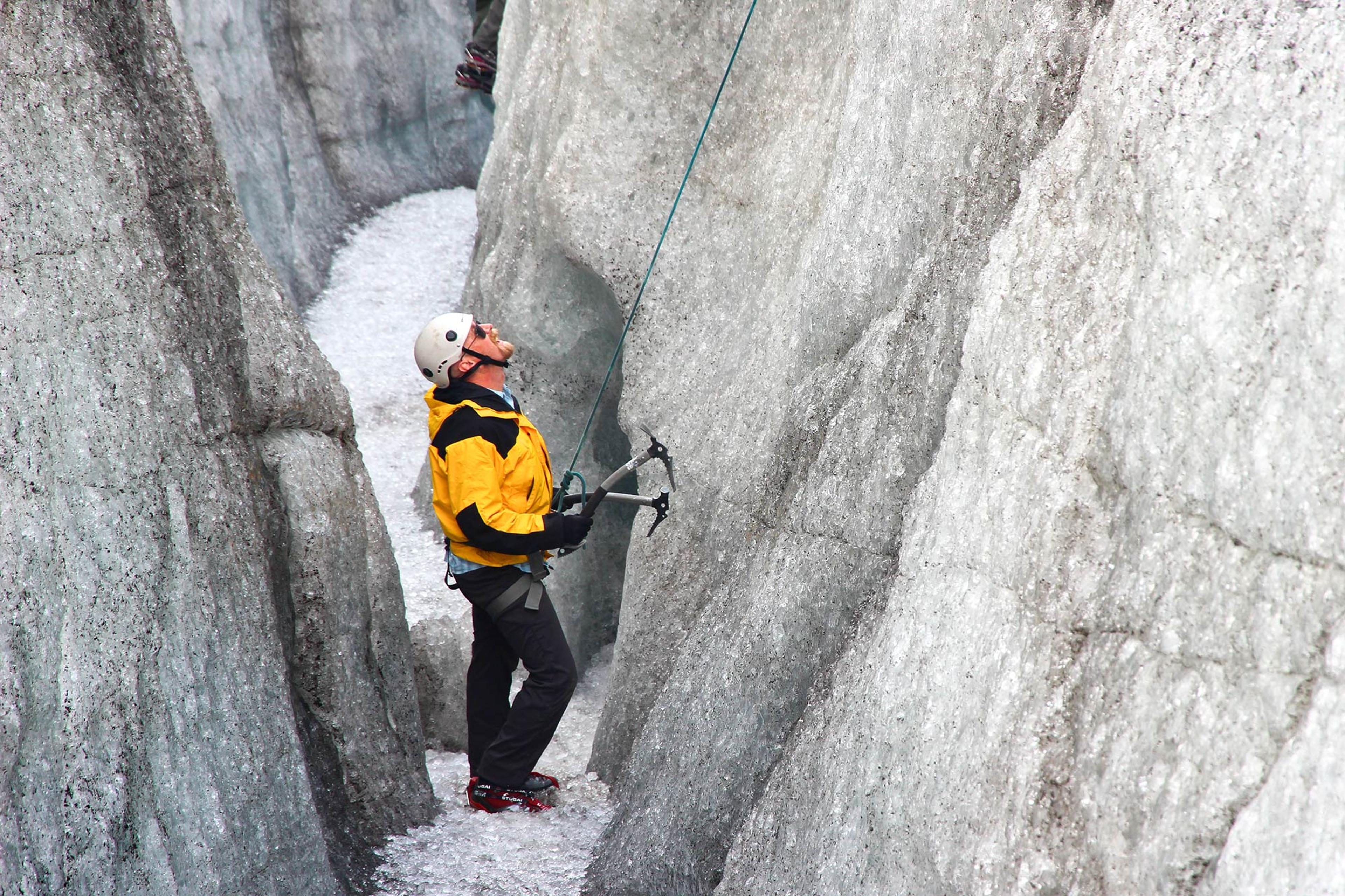 A climber in a yellow jacket prepares to ascend a glacier crevasse, gripping his ice axes tightly against the icy walls.