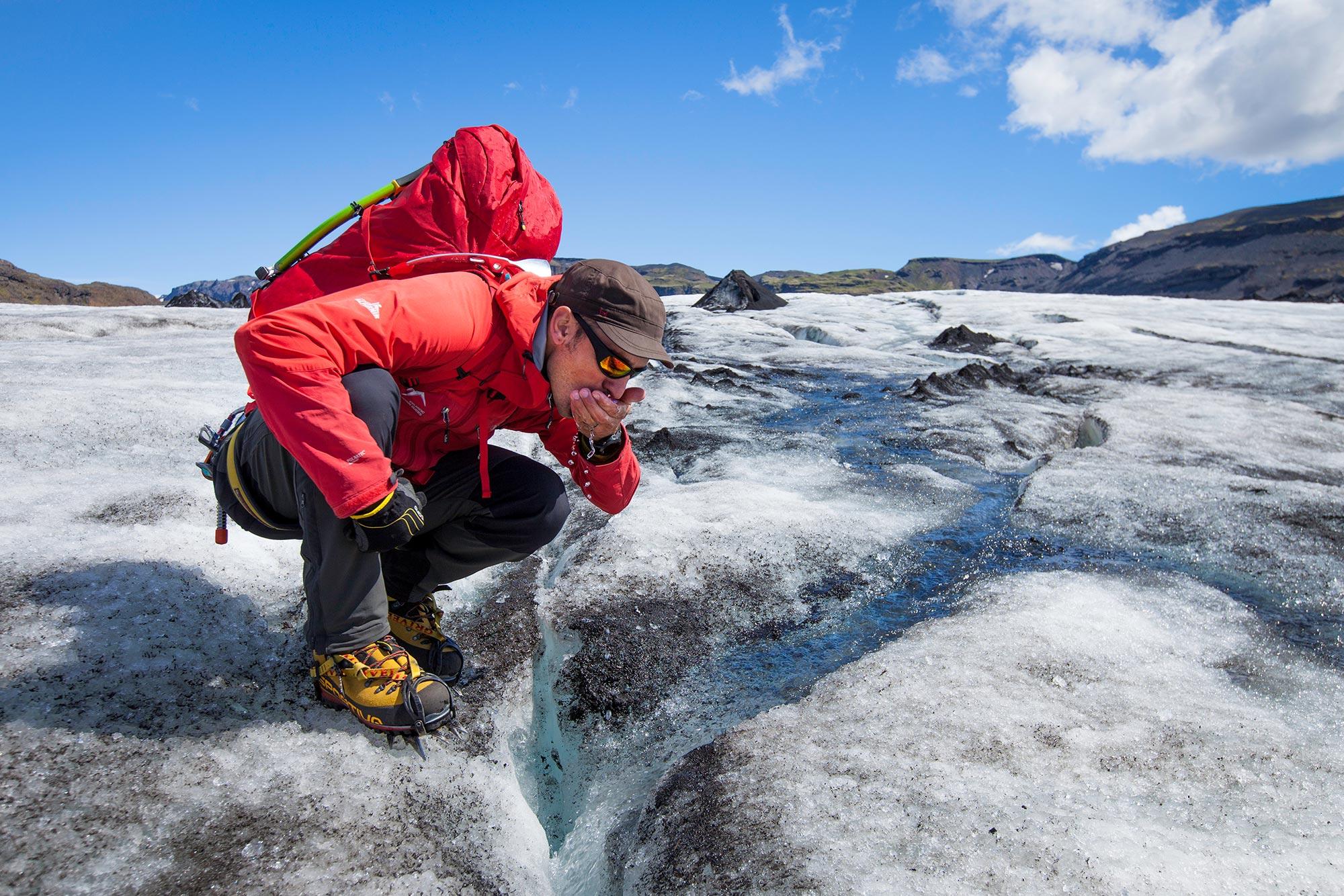 Man in red standing on a glacier drinks water from a creek on the glacier