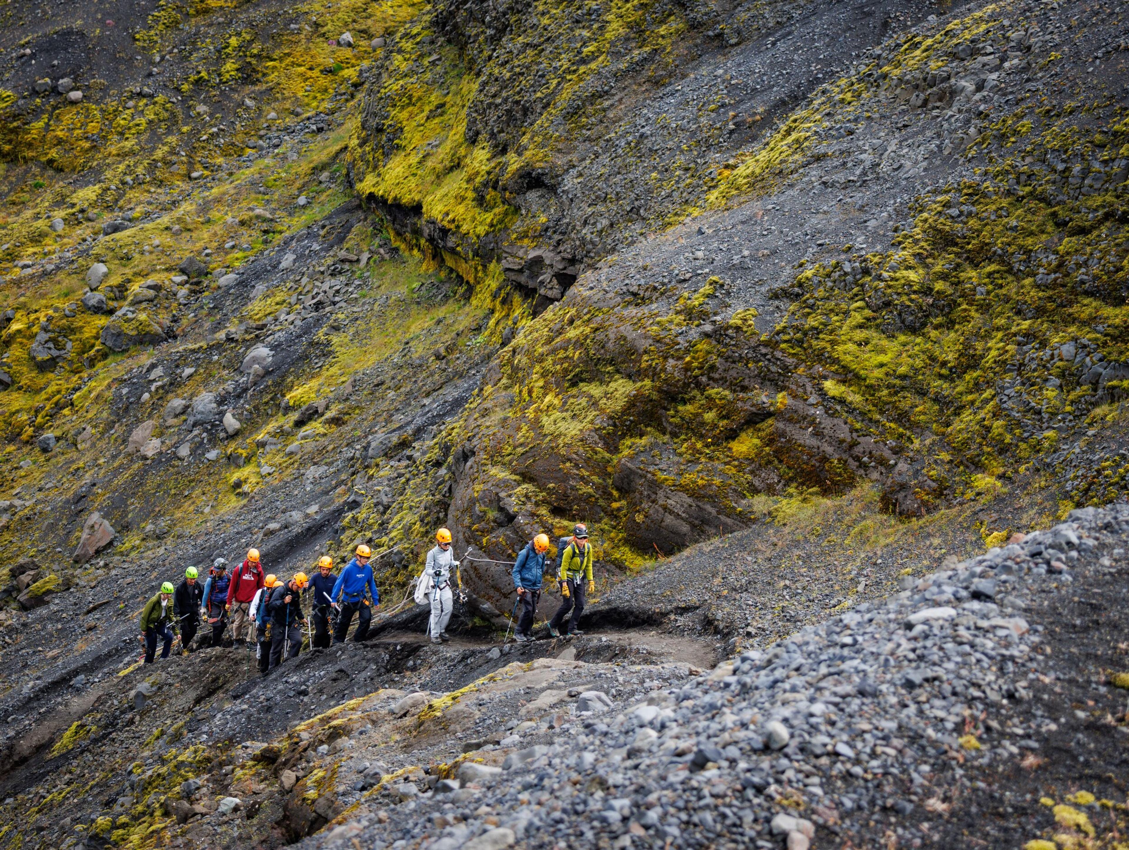 A group of hikers in colorful helmets makes their way along a narrow trail on a steep slope streaked with vivid yellow sulfur deposits.