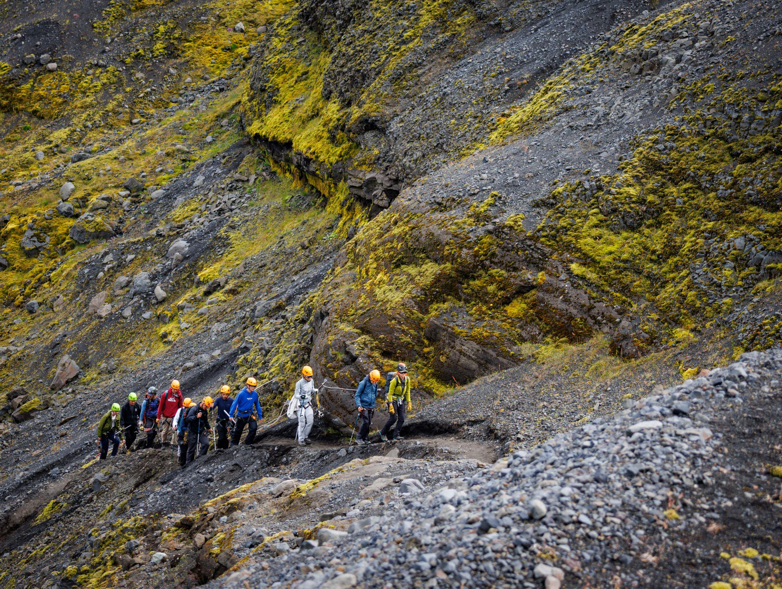 A group of hikers in colorful helmets makes their way along a narrow trail on a steep slope streaked with vivid yellow sulfur deposits.