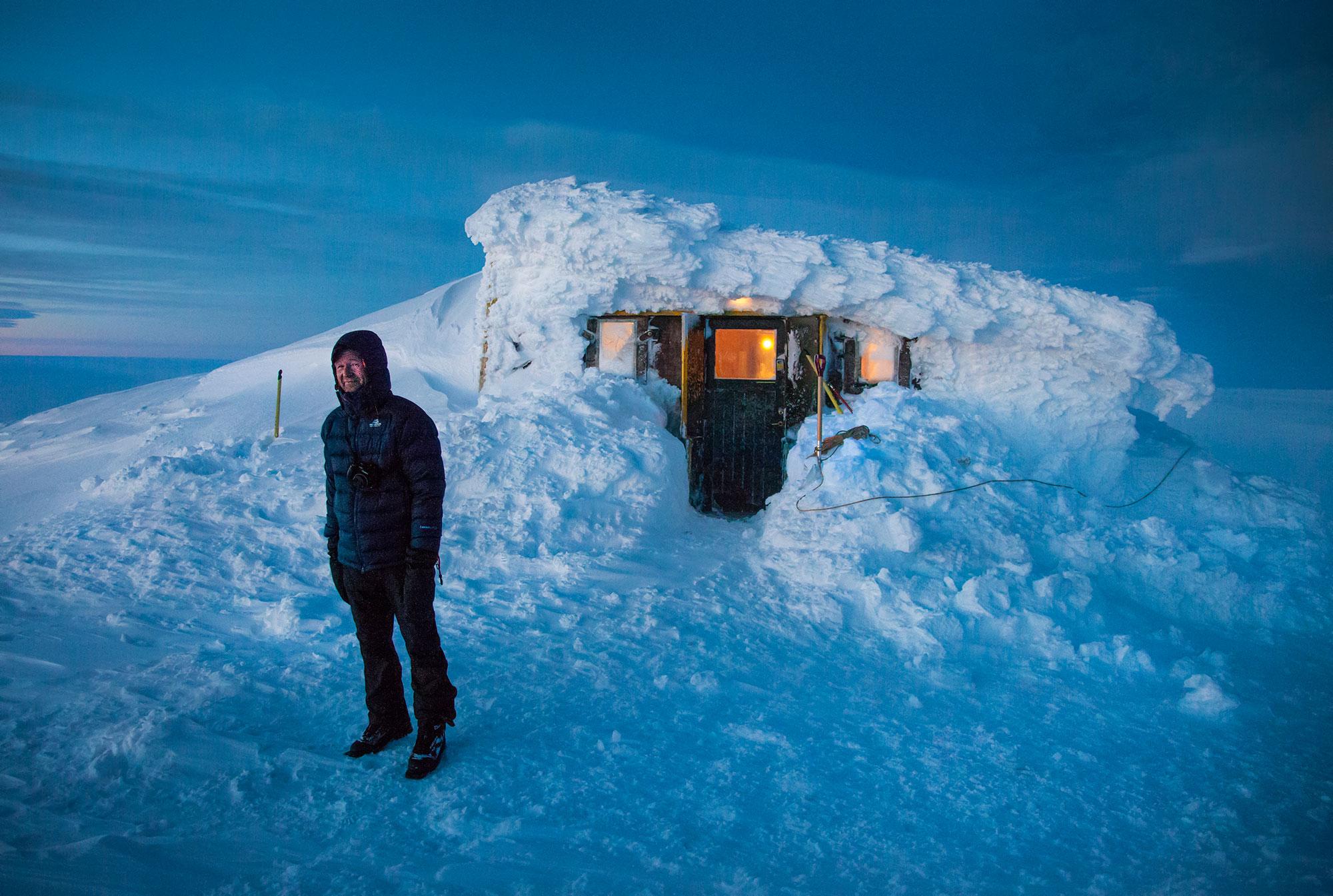 A man standing in front of snow covered Grímsfjall mountain hunting the evening so the lights of the hut are very visible shining through the snow
