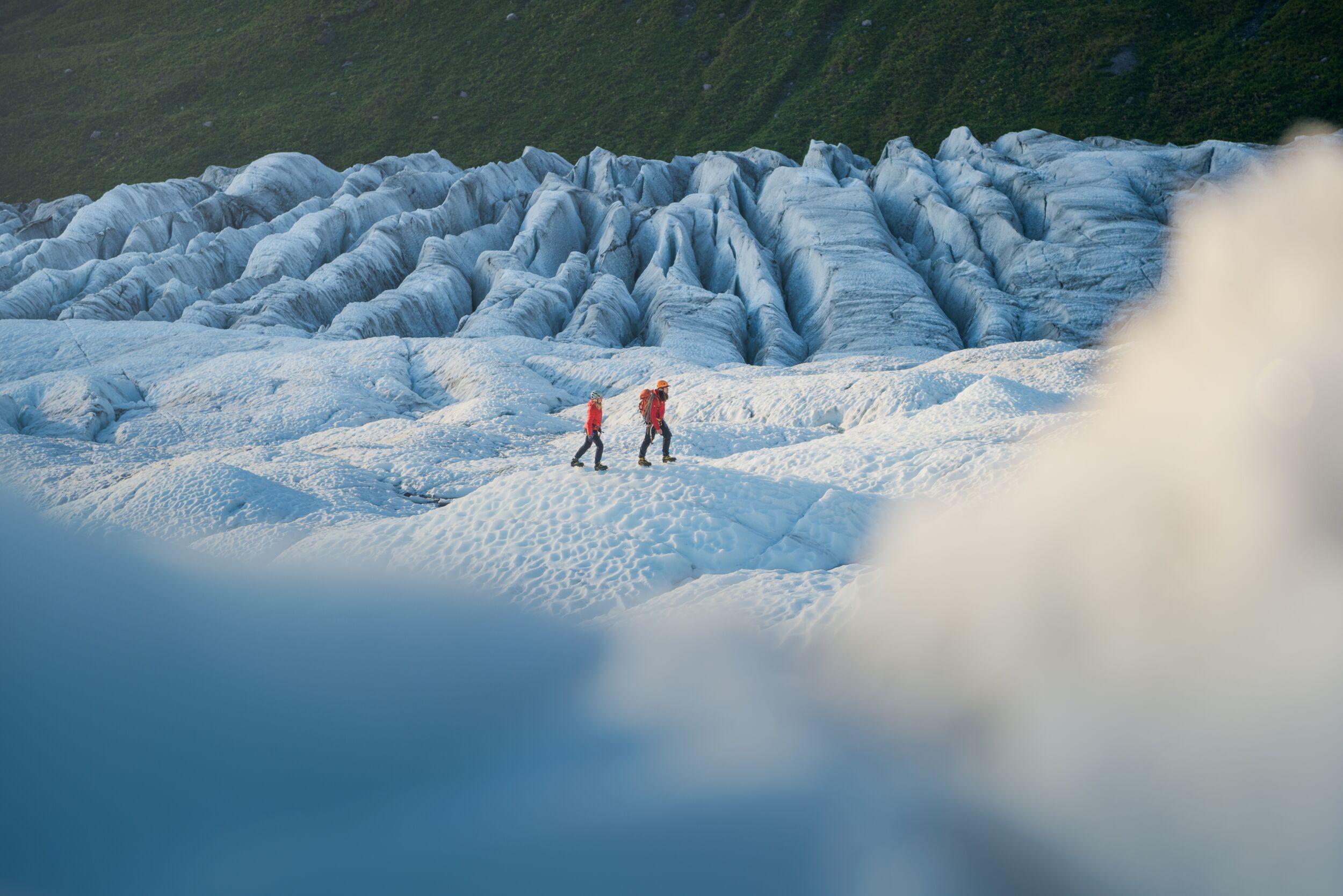 "Two hikers in red jackets walk across a rippled glacier surface, framed by an ice foreground and rows of textured blue-white ice ridges behind them