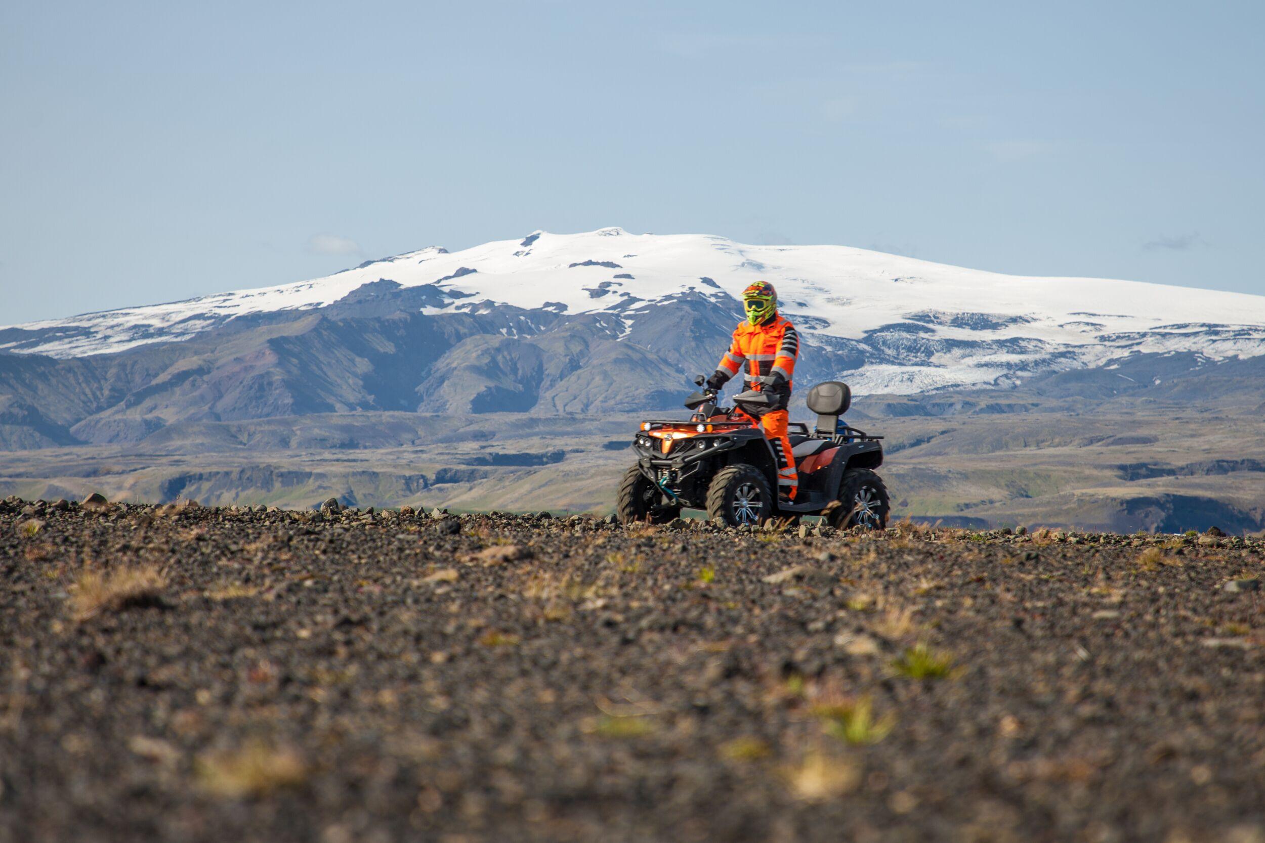 A rider in bright orange gear navigates an ATV across Iceland’s rocky terrain, with a stunning snow-capped mountain in the background.