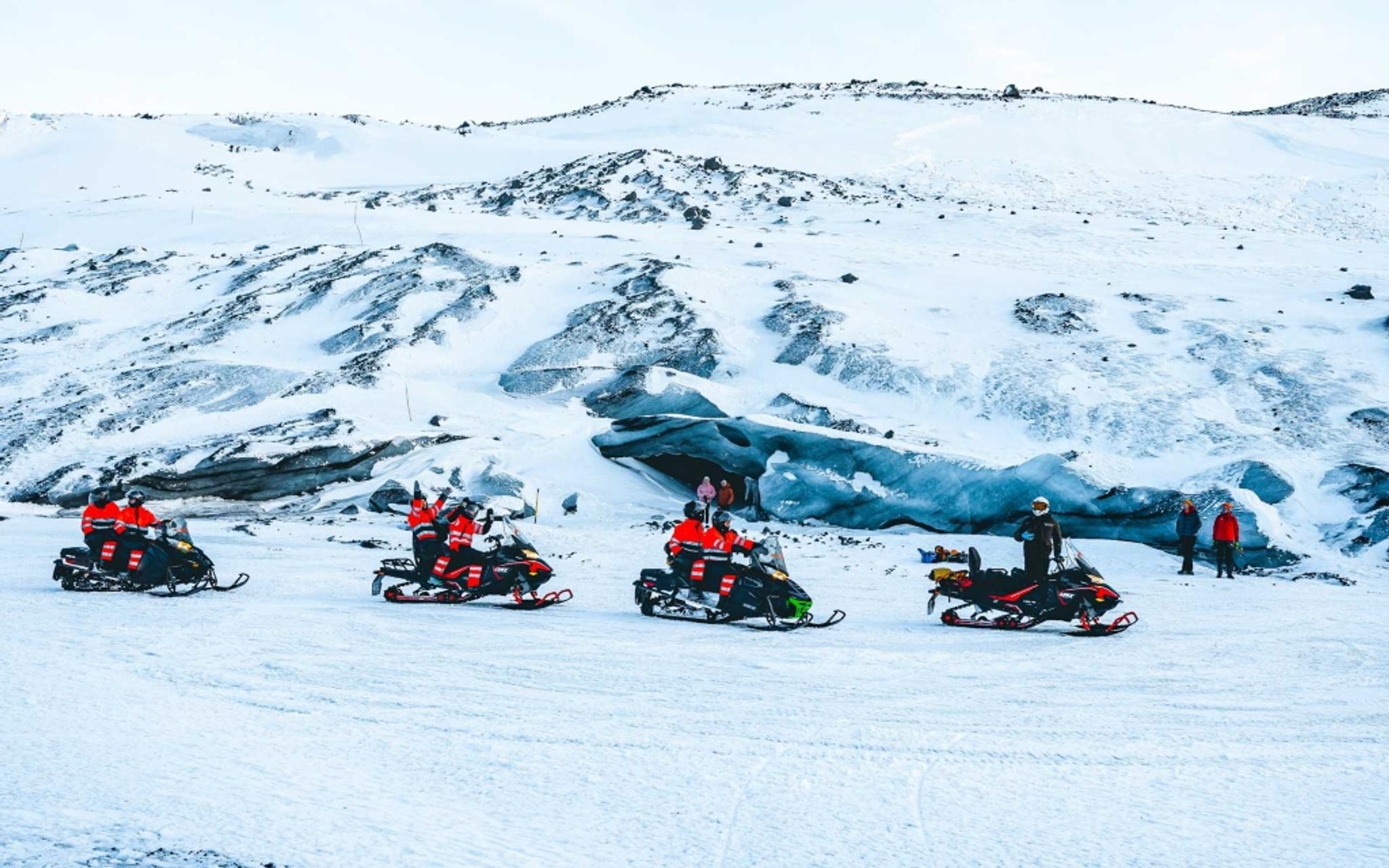 A convoy of snowmobiles riding through the snow with blue ice walls in the background
