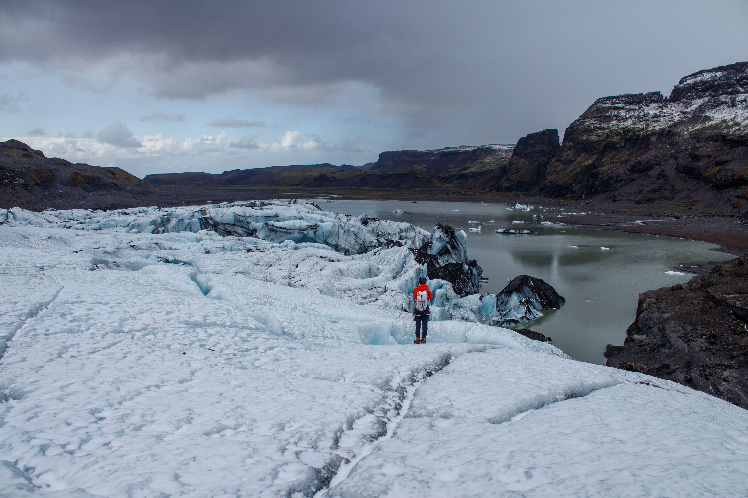 Person wearing a red backpack standing on Sólheimajökull glacier, surrounded by rugged mountains, icy terrain, and a glacial lagoon under a dramatic sky.