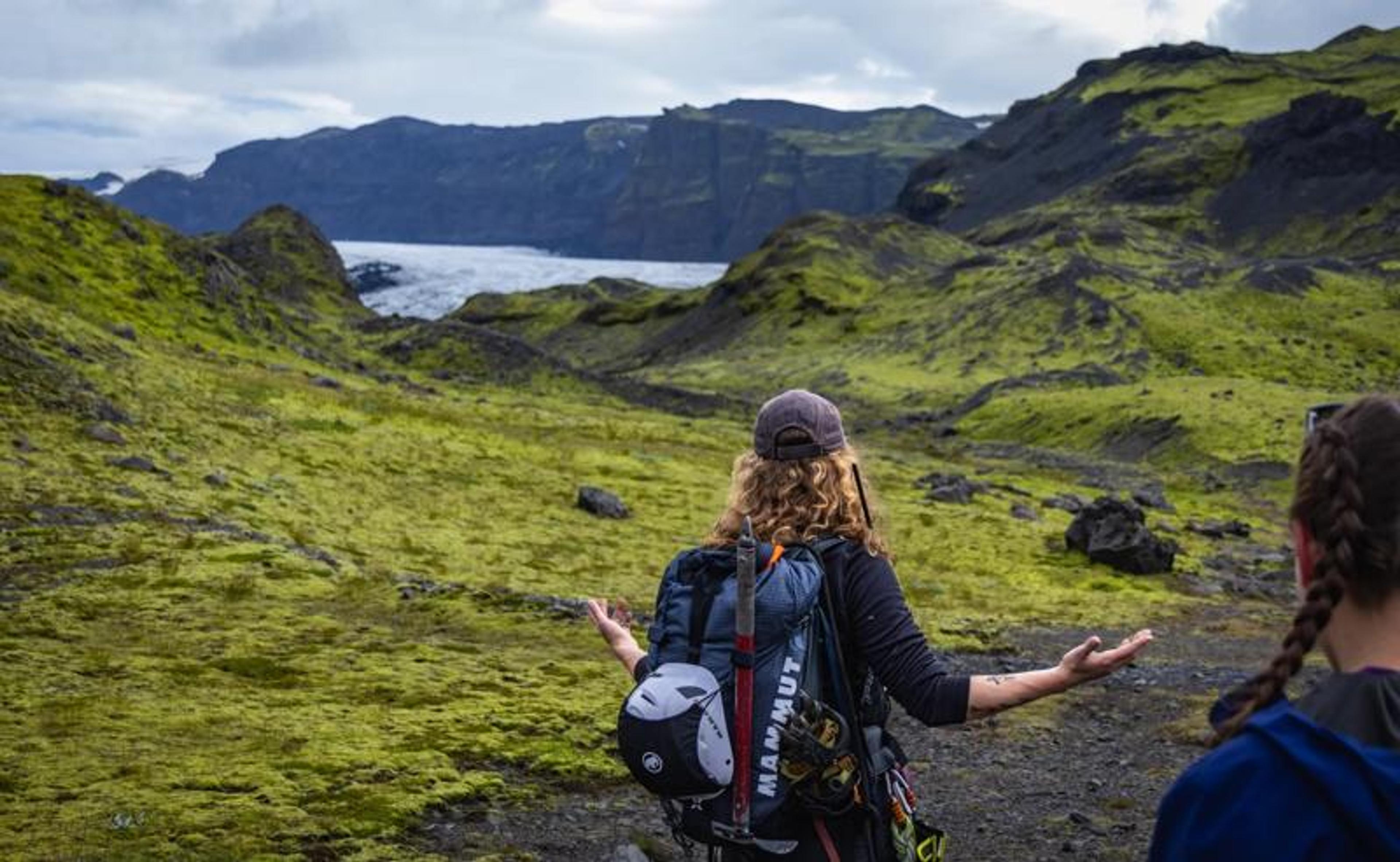 Hikers with backpacks and a climbing helmet walk through a moss-covered volcanic landscape, gesturing toward dramatic sea cliffs and a distant glacier tongue meeting the ocean.