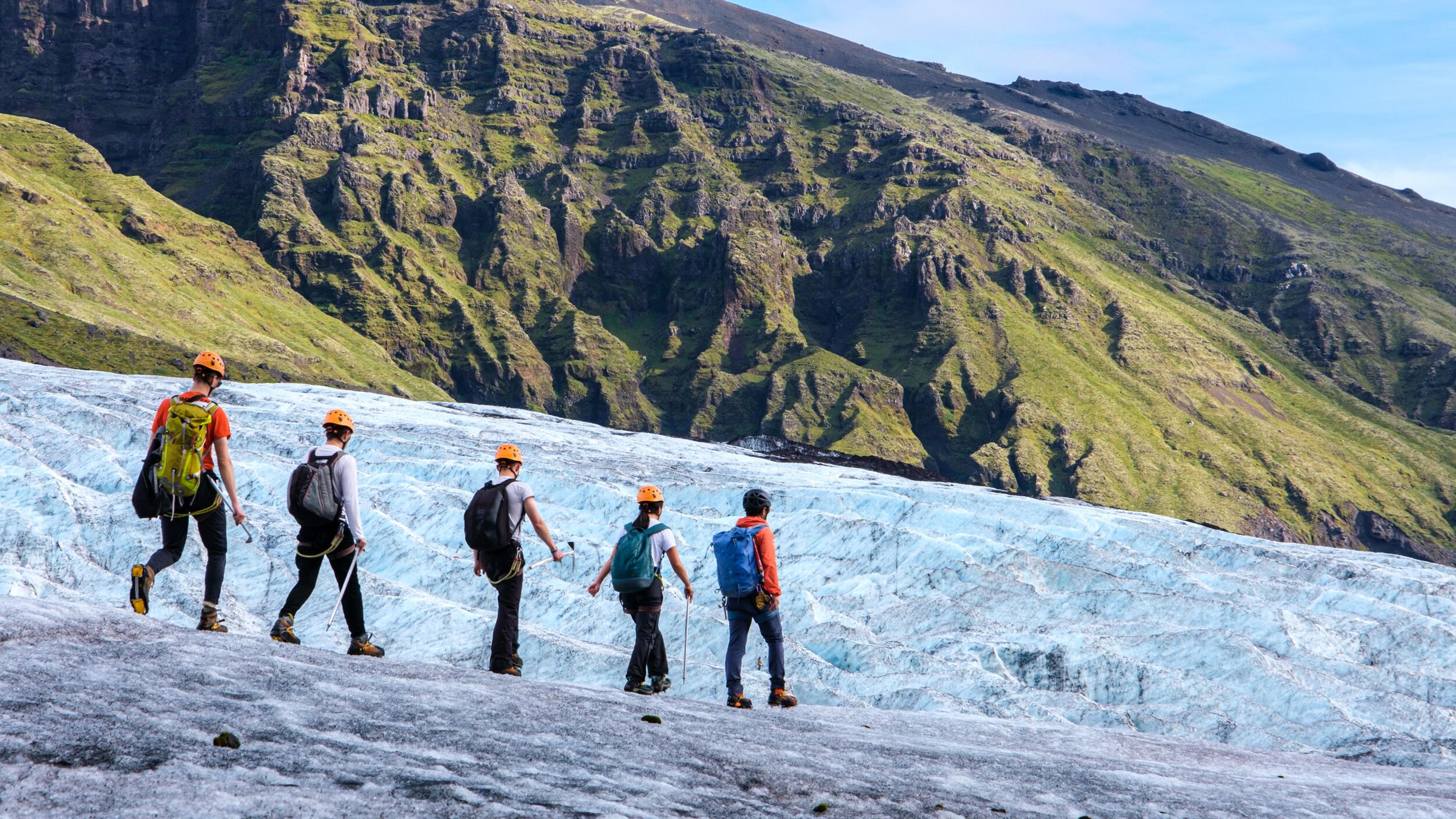 A group of hikers equipped with helmets and crampons traverses a glacier, with towering green cliffs providing a dramatic backdrop.