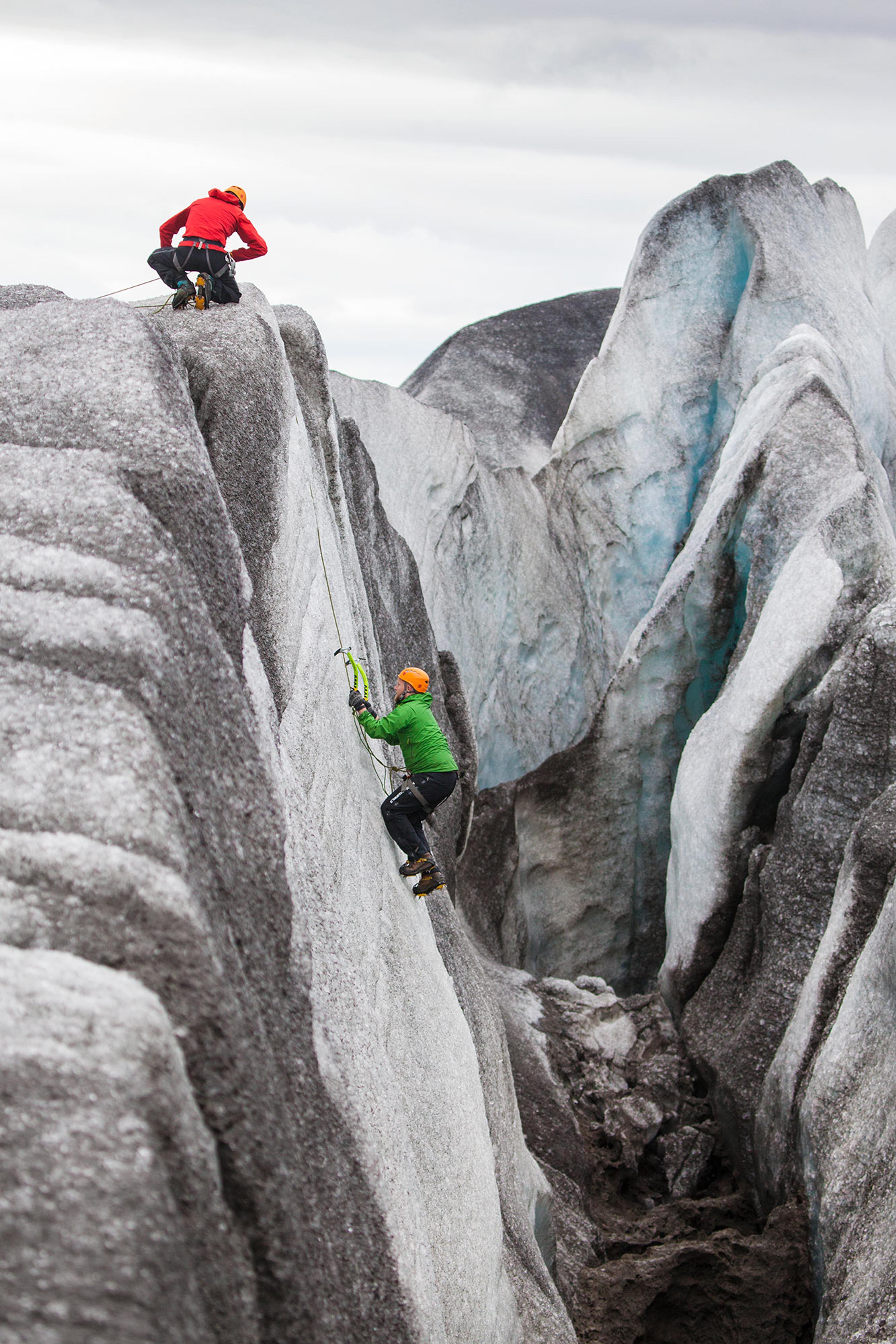 Two climbers celebrate at the summit of a glacier ridge, holding ice axes and wearing bright gear, surrounded by icy terrain.