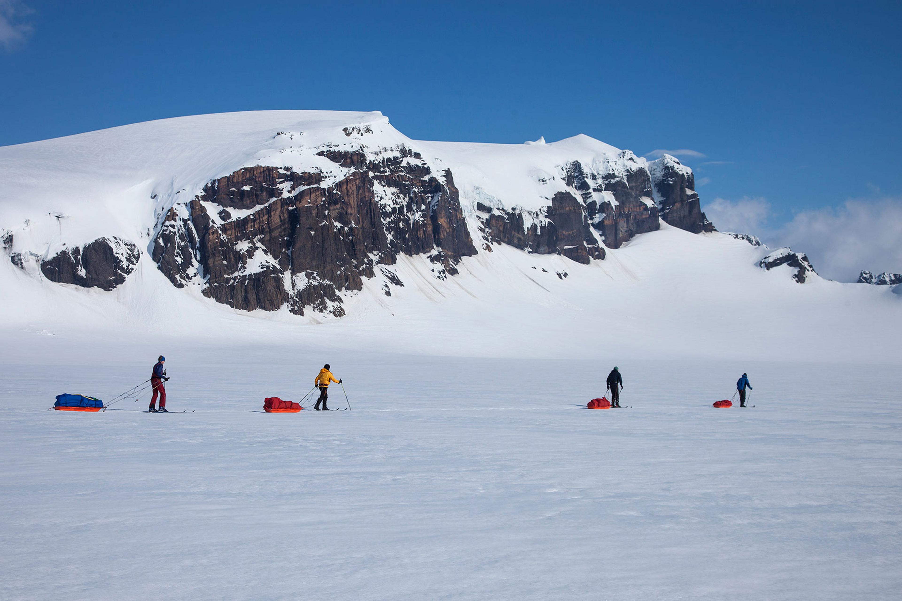 Four persons skiing by a range of mountains and dragging pulkas behind