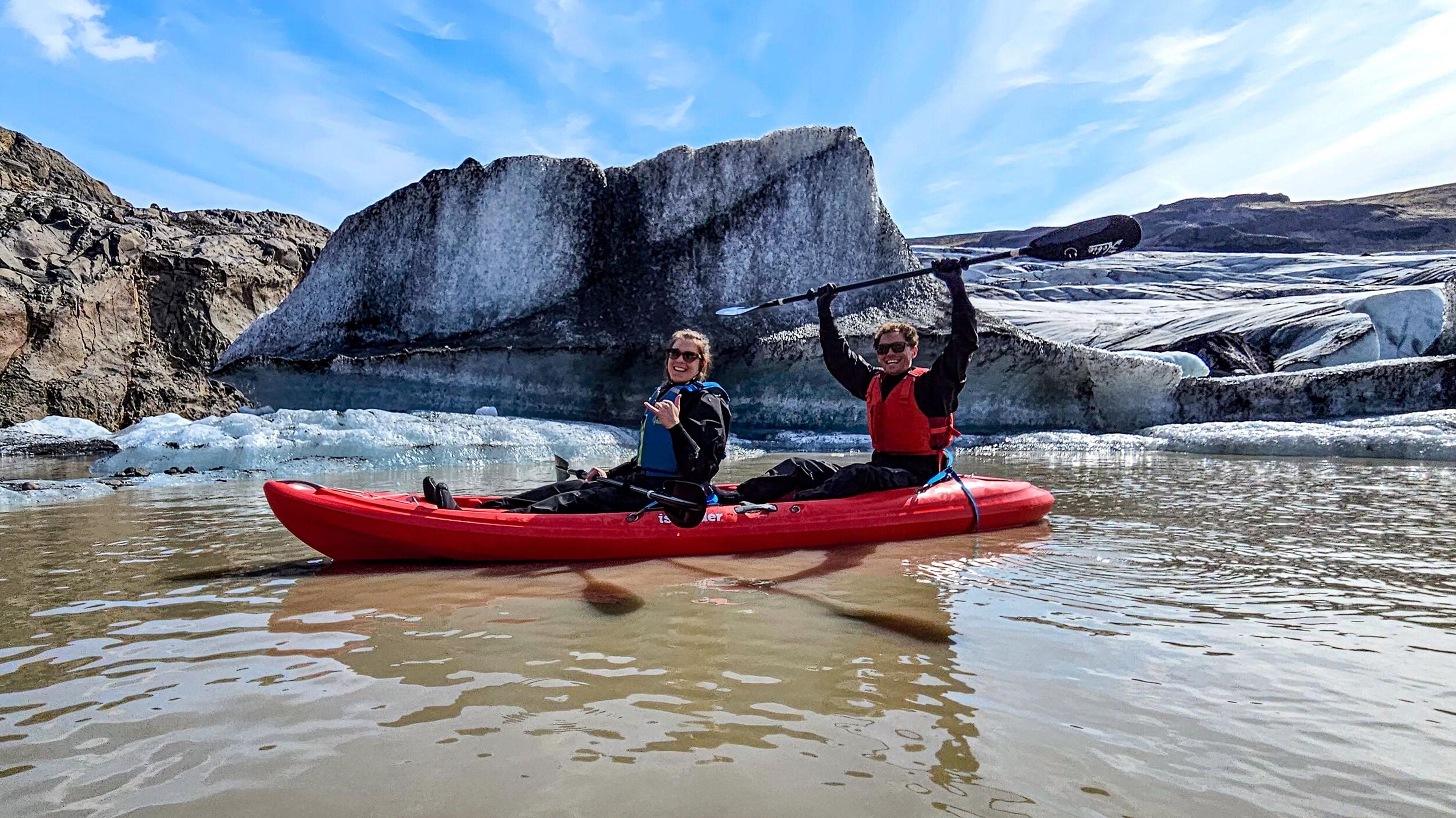 Two people in a red tandem kayak pose on a glacial lake, one raising a paddle overhead, with towering ice formations and a glacier behind them under a bright blue sky.