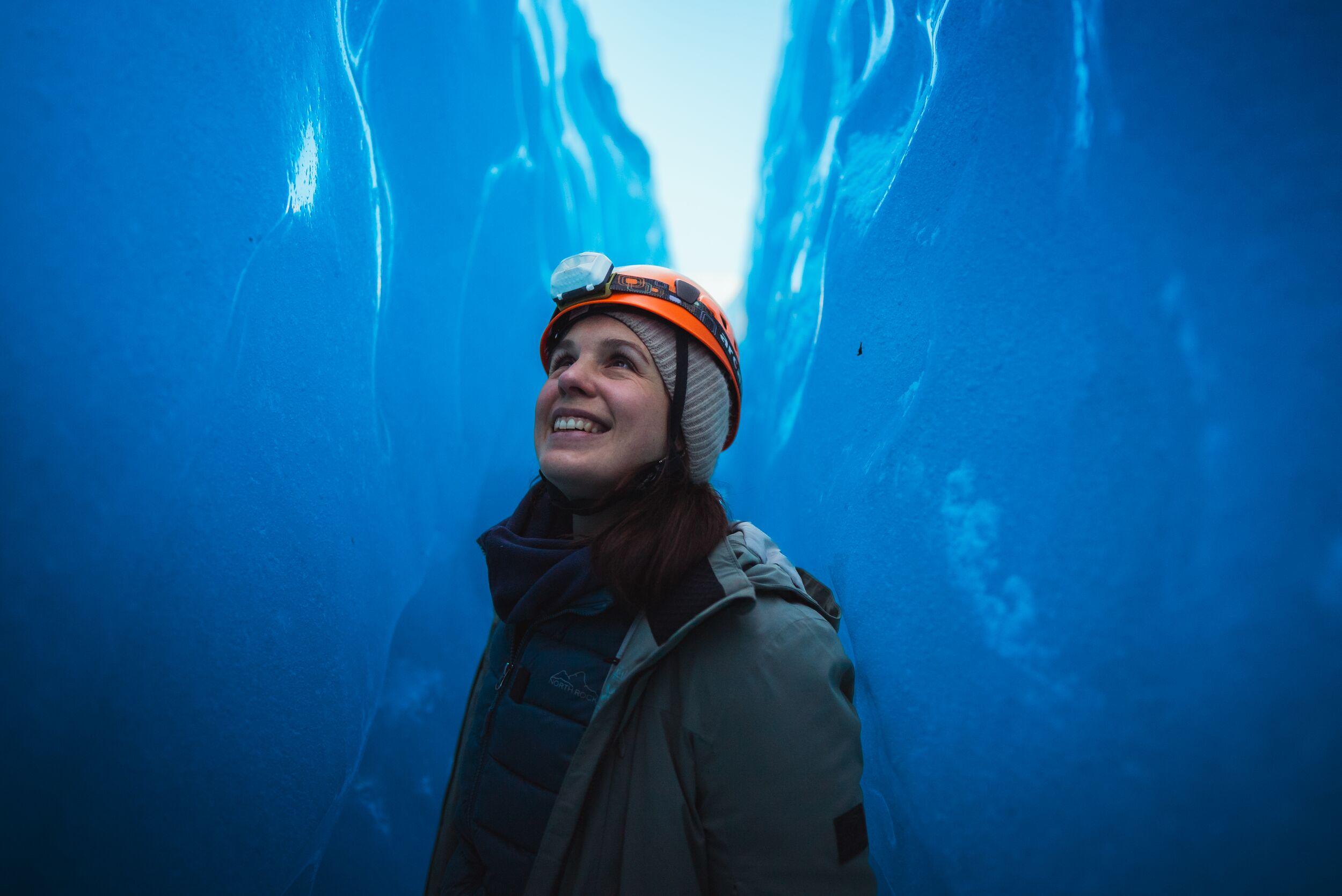 A woman with a helmet stood in a dark blue crevasse looking up and smiling