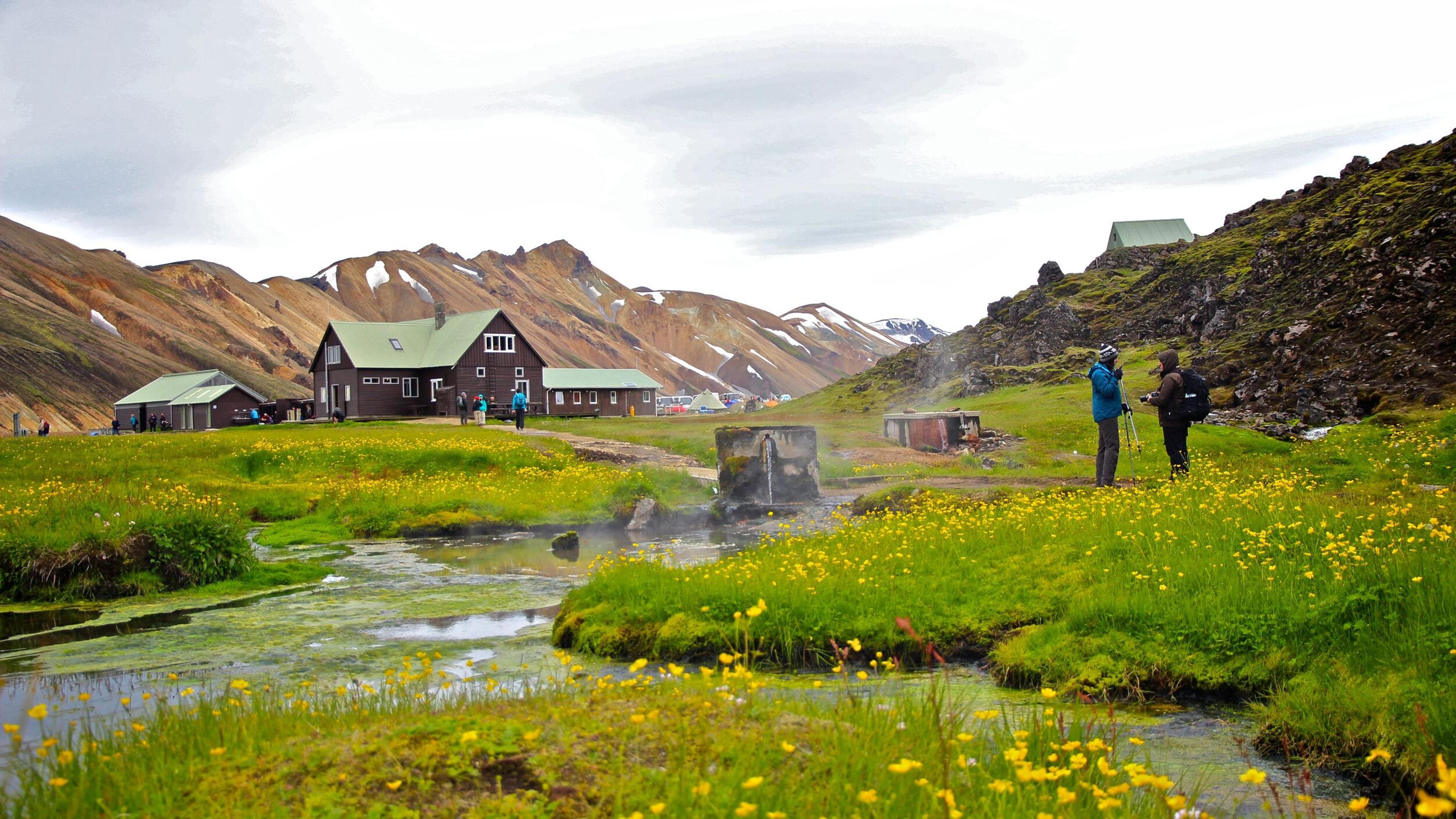 Expansive view of Landmannalaugar’s volcanic terrain, with dark lava fields, patches of snow, and distant mountains under a cloudy sky.