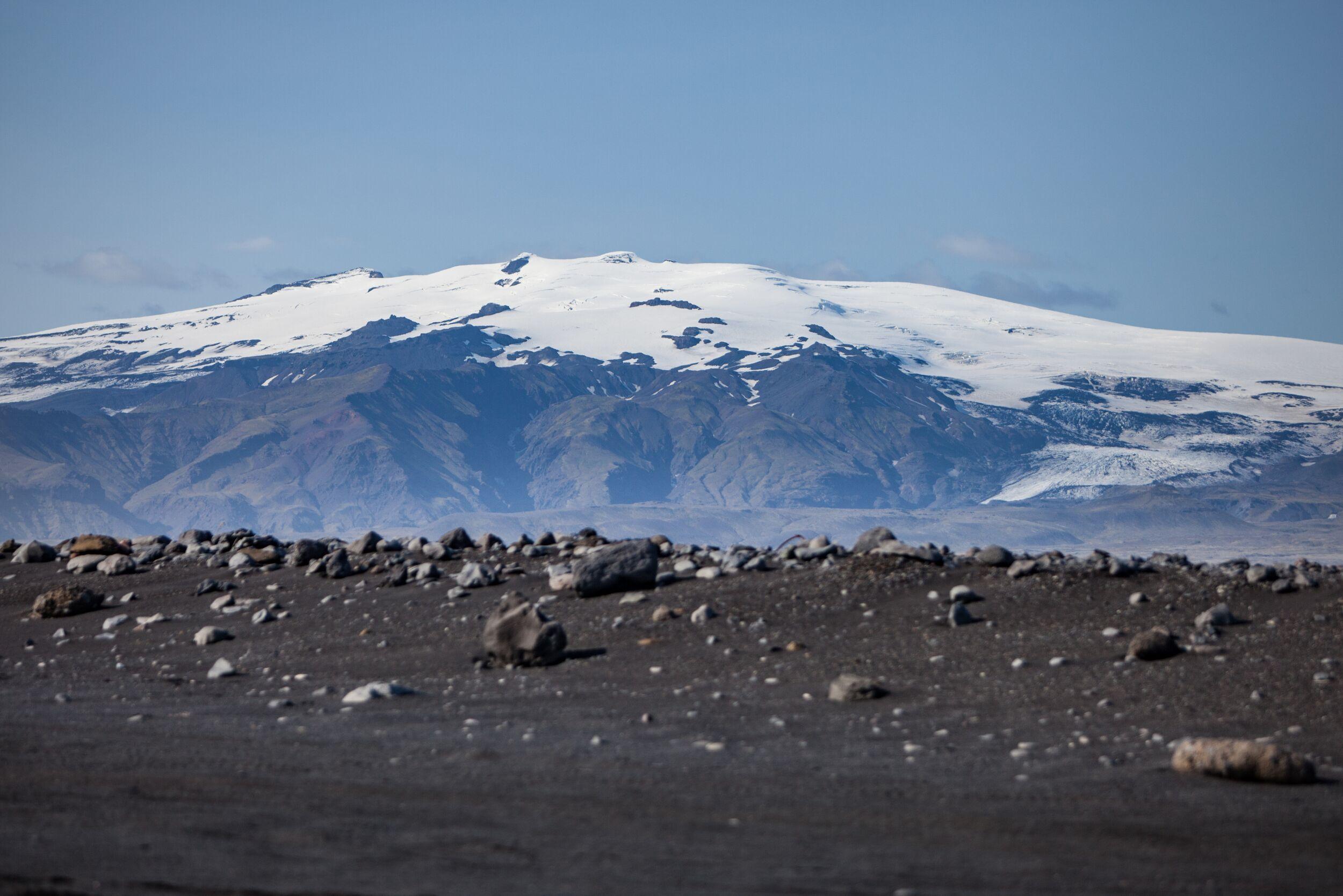 A snow-capped volcano rises majestically above a rugged landscape of black sand and scattered rocks.