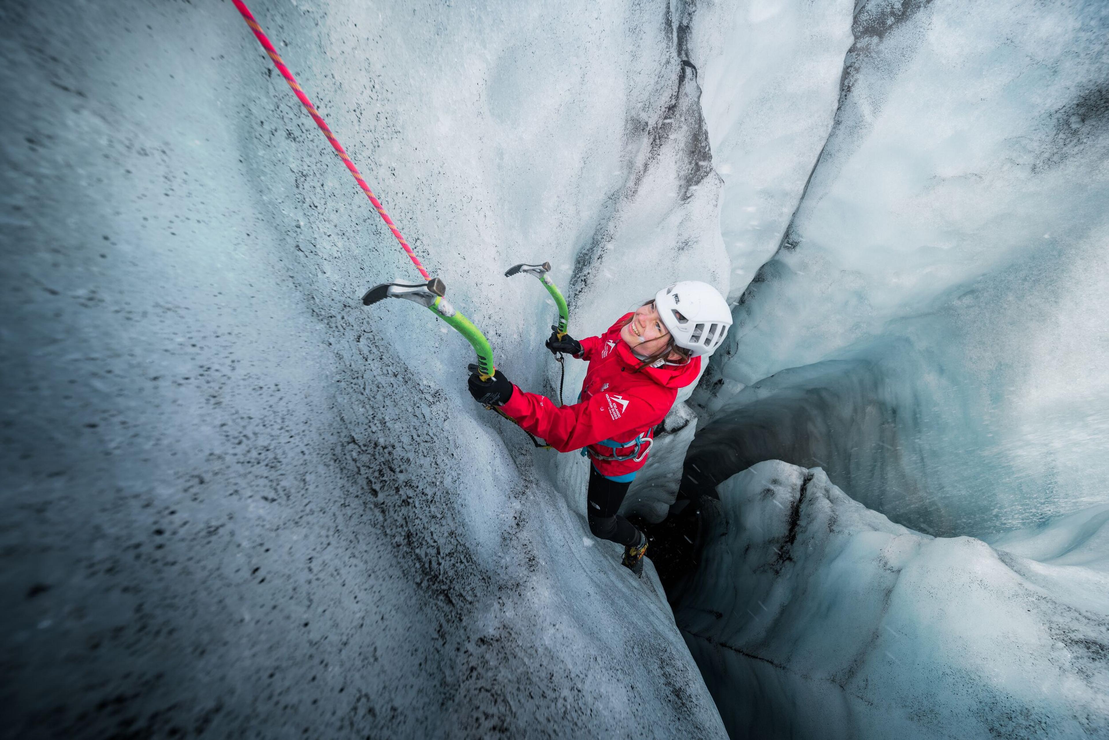 A climber in a red jacket and helmet uses ice axes to ascend a steep, icy wall in a glacier crevasse, highlighting the thrill of ice climbing in Iceland.