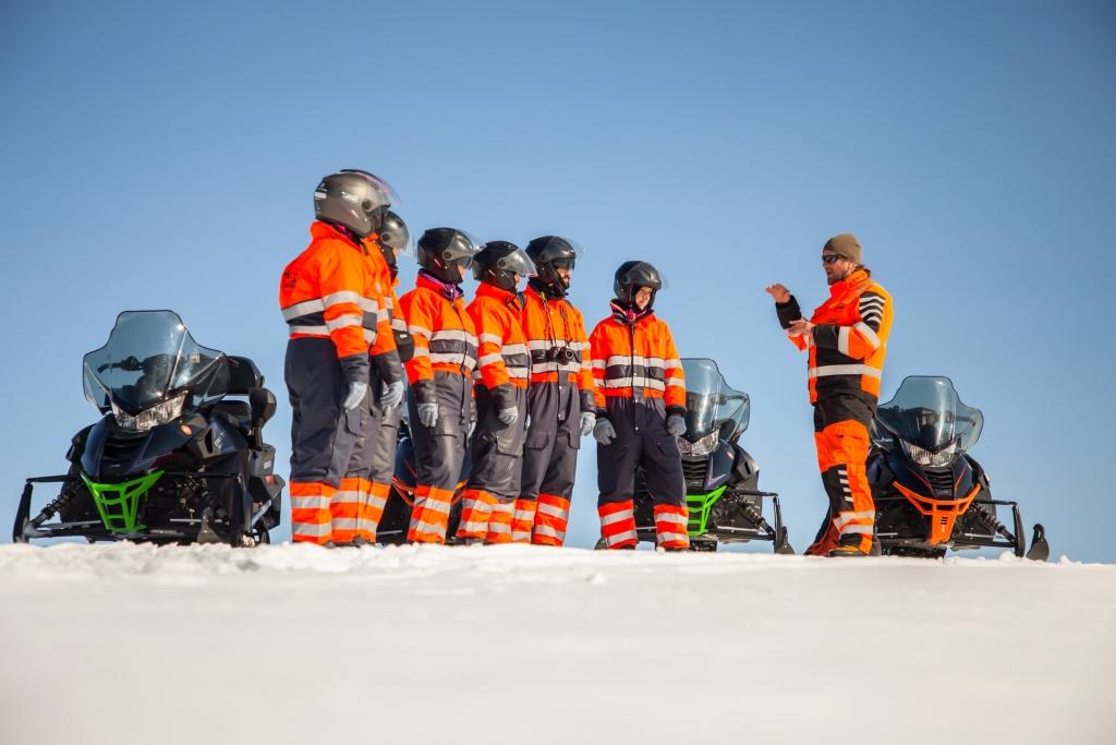 People in orange gear with snowmobiles taking a break on a snowy hillside.
