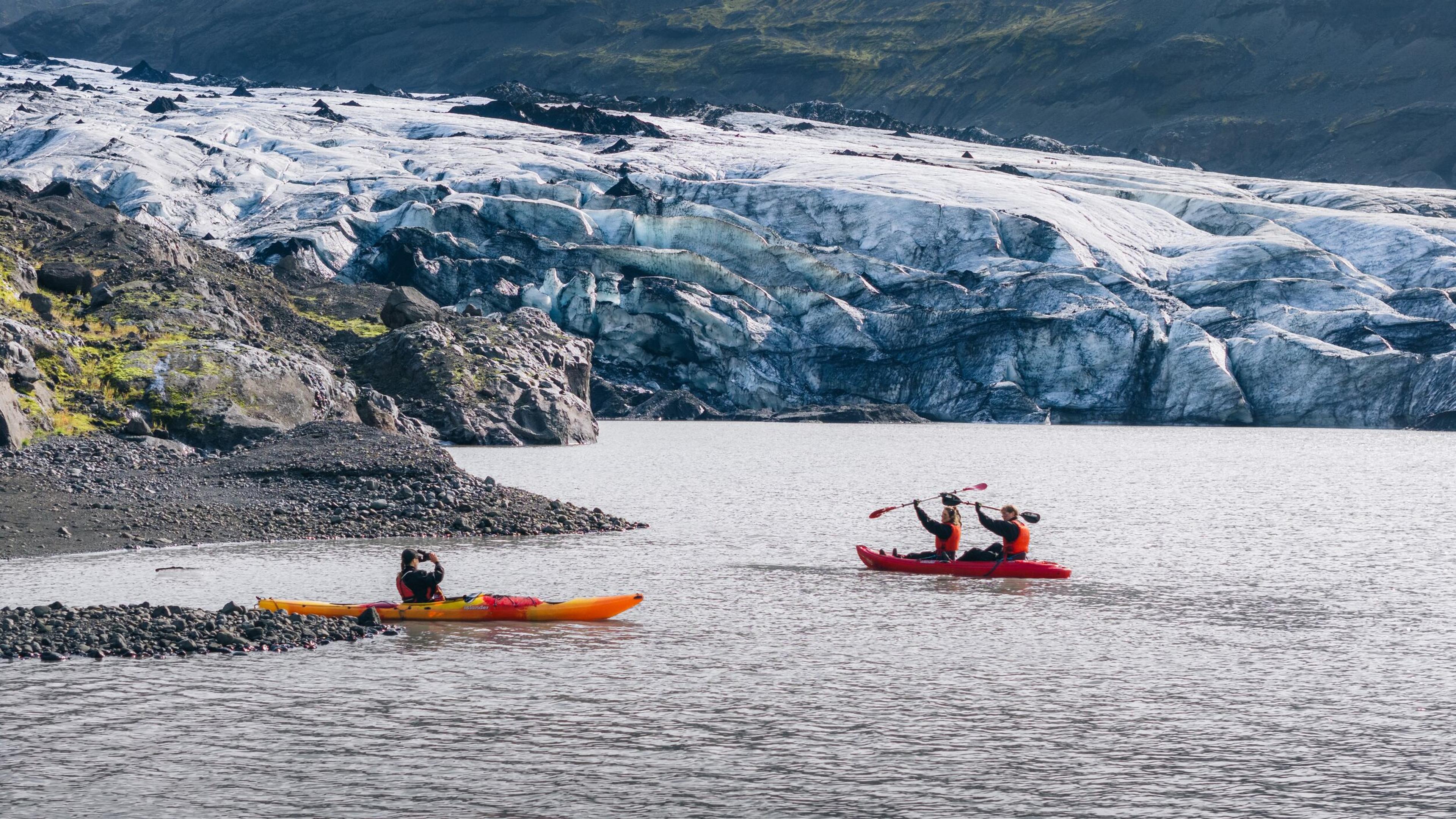 Two kayaks carrying people in life jackets paddle across a glacial lake, with a vast, rugged blue-white glacier and rocky shoreline behind them.